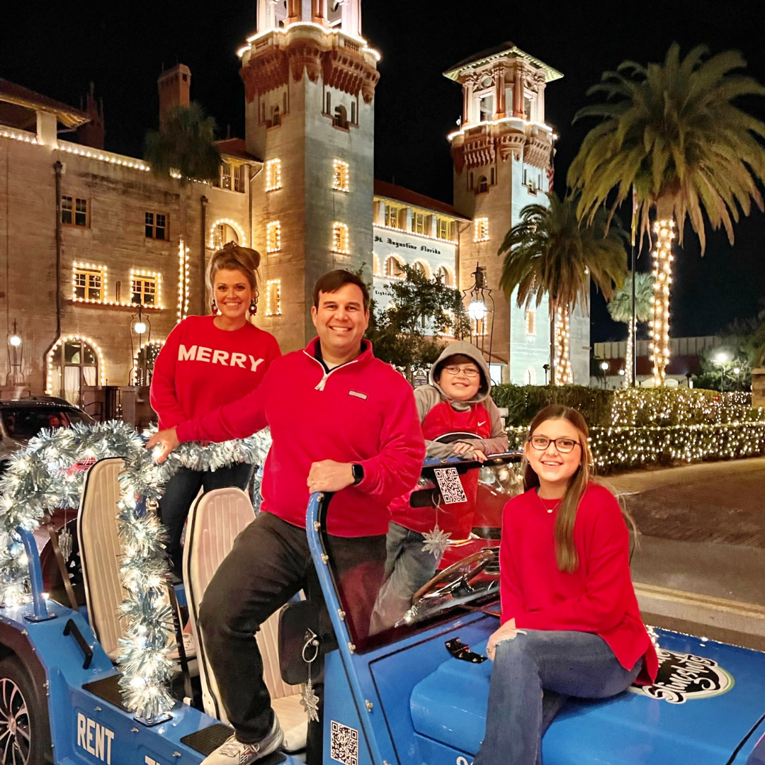 Family in red sweaters smiles in a decorated blue car at night; building with Christmas lights in background.