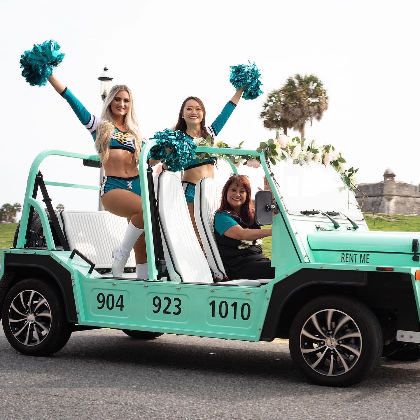 Three cheerleaders posing in a mint-green Jeep decorated for a parade on a street.