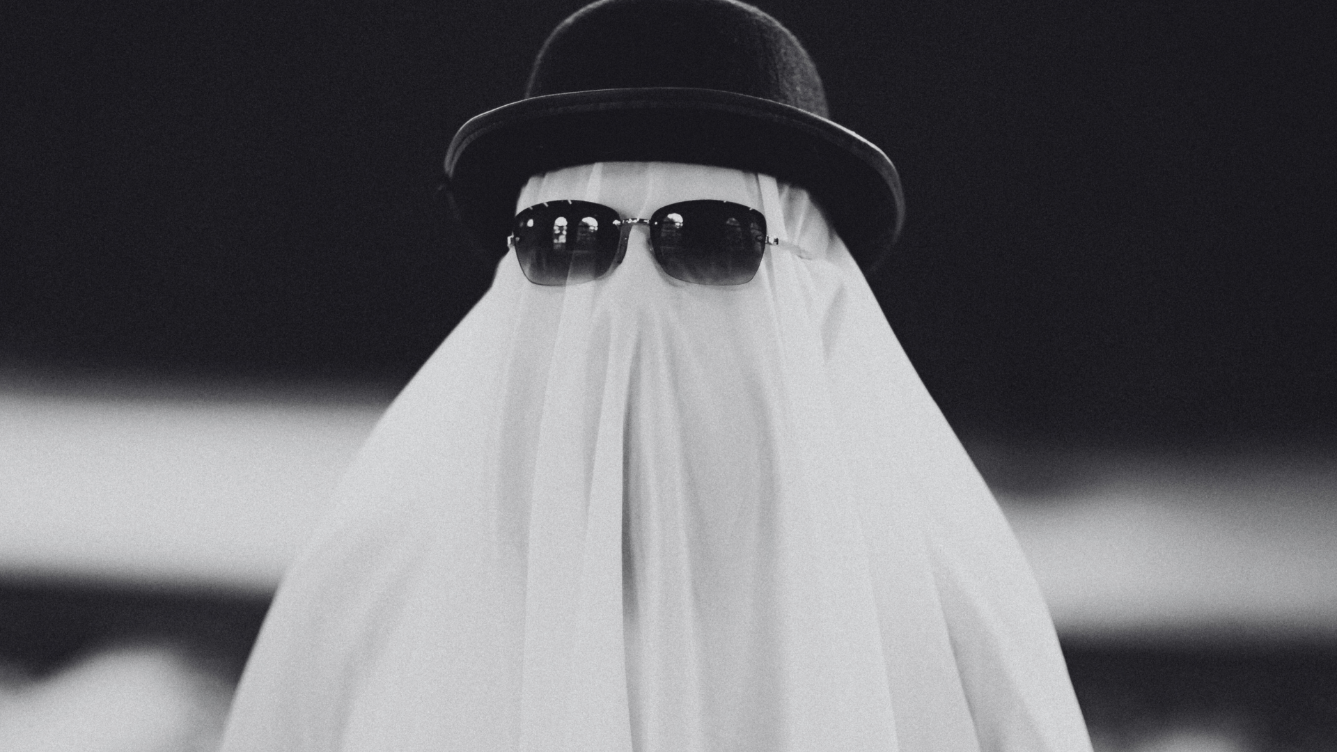 Person wearing sunglasses and a dark hat, with a white ghost-like veil covering the body