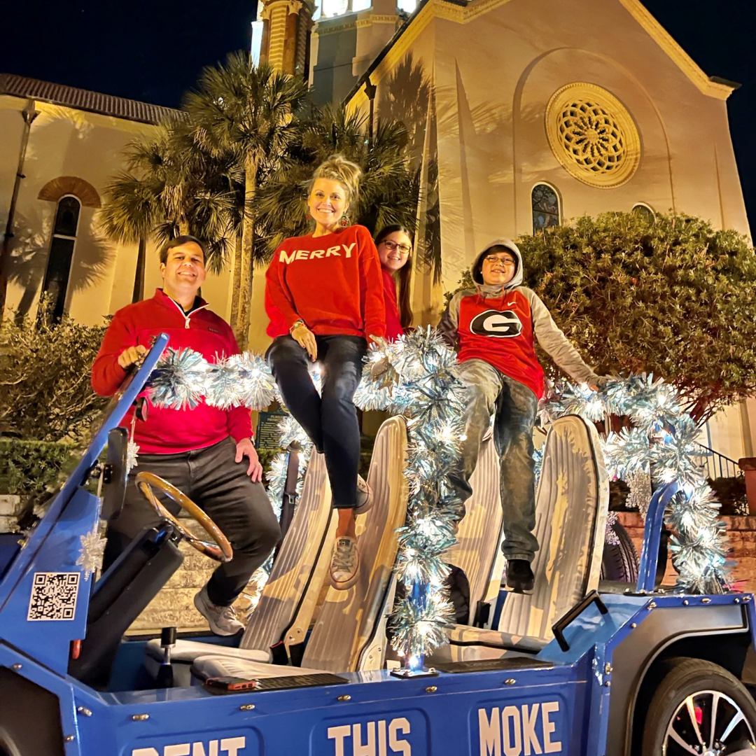 Family in red sweaters on a decorated blue Moke car in front of a building at night for St. Augustine Nights of Lights tour.