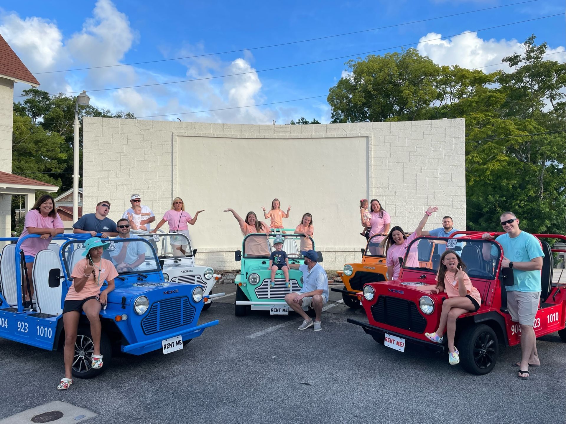 Group poses with colorful mini moke Jeeps in front of a white wall. Some people are waving.