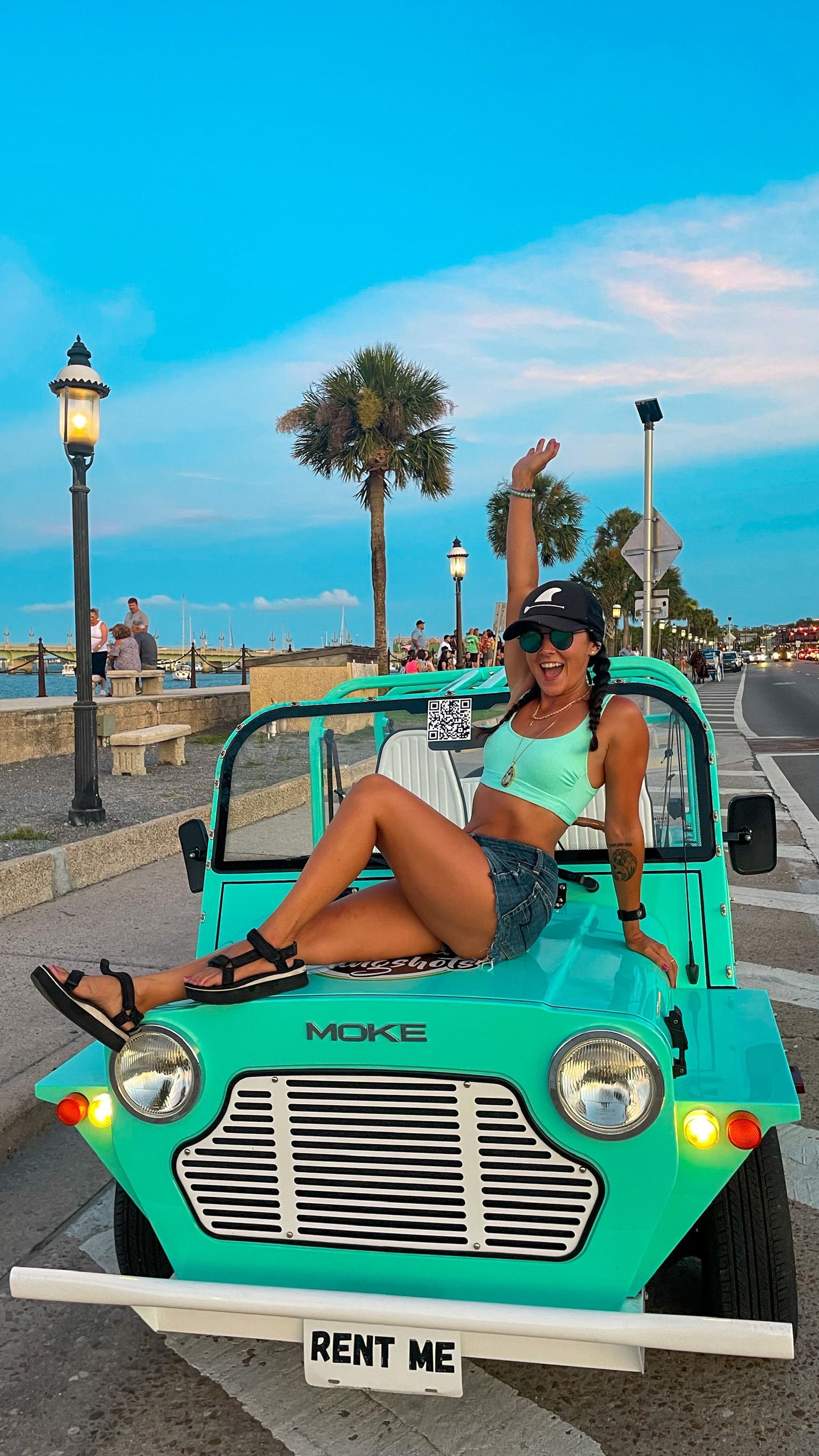 Person lounging in a turquoise RENT ME car by the waterfront at sunset