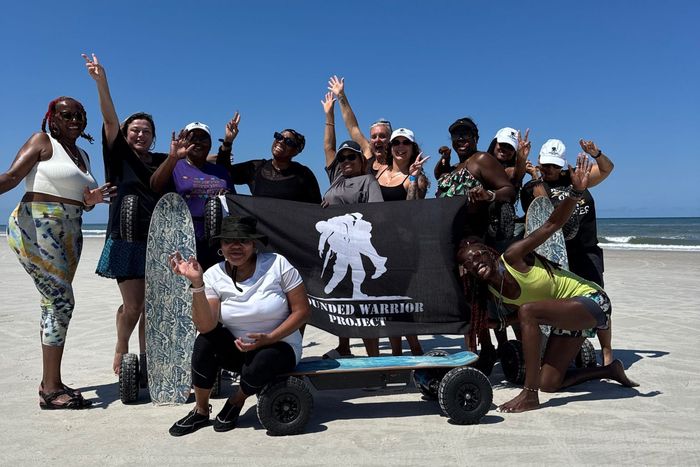 Group of people posing on a beach with a black banner, many waving and smiling under a blue sky