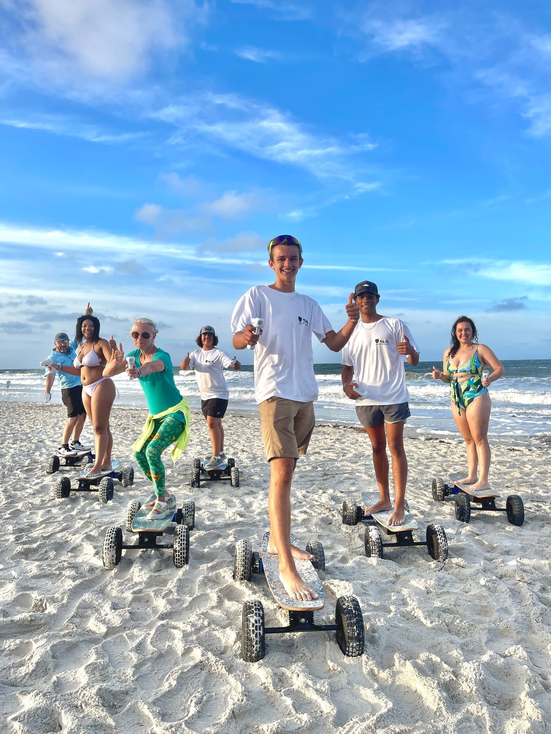 Six people jogging on all-terrain roller skates across a sandy beach under a blue sky.