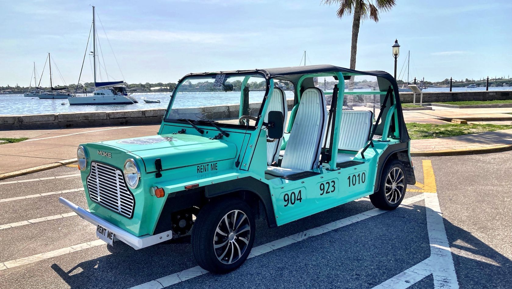 Turquoise vintage-style electric shuttle parked by a marina, with sailboats and palm trees in the background