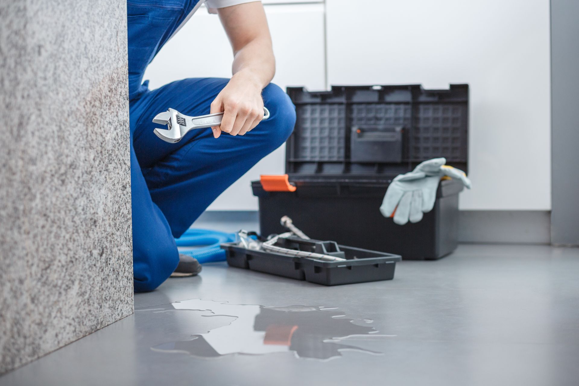 a person in blue overalls kneeling on a wet floor holding an adjustable wrench.