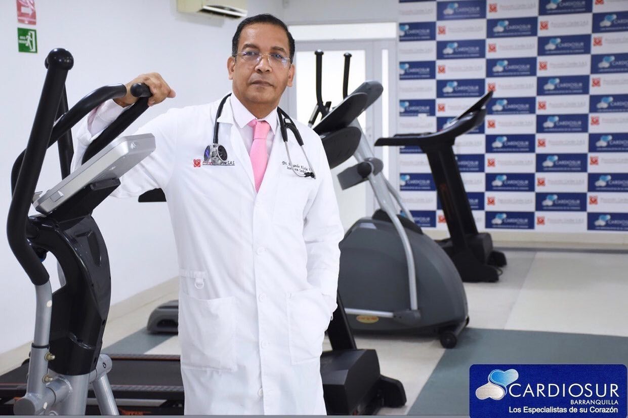 Médico con bata blanca de pie en un gimnasio junto a máquinas de ejercicio, con un fondo de pared