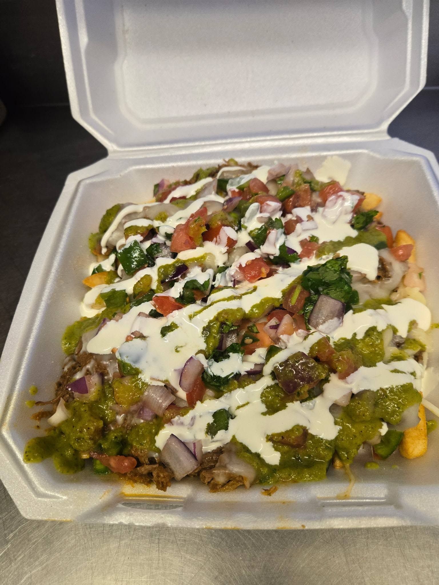 Takeout container of loaded nachos topped with cheese, sauce, onions, and herbs