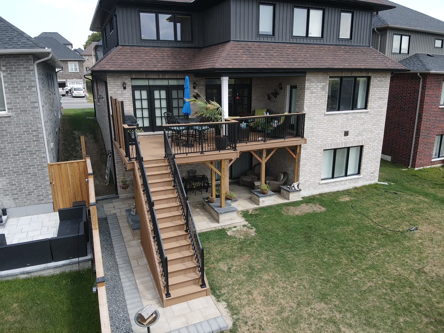 Two-story house with wooden deck and stairs. Beige brick exterior, dark roof, black railings, and a green lawn.