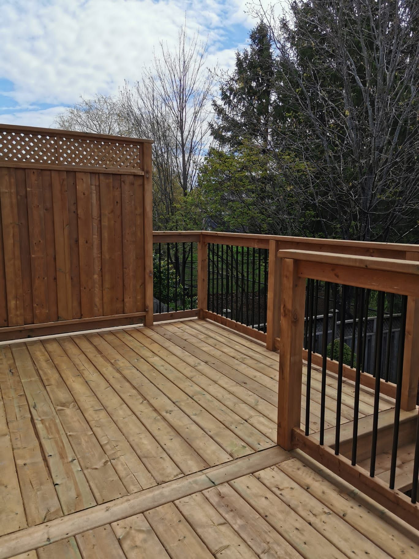 Wooden deck with railings and privacy fence, surrounded by trees under a cloudy sky.