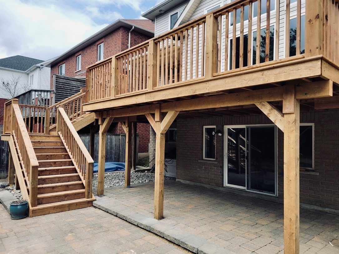 Wooden two-story deck with stairs leading down to a stone patio. Brick house in the background.