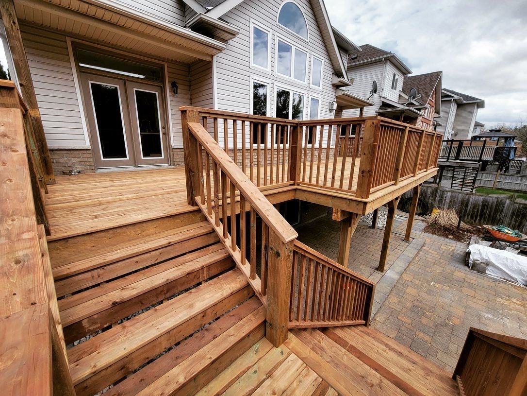 Wooden deck with stairs leading to a house, brown wood railing, stone facade, and paved patio.
