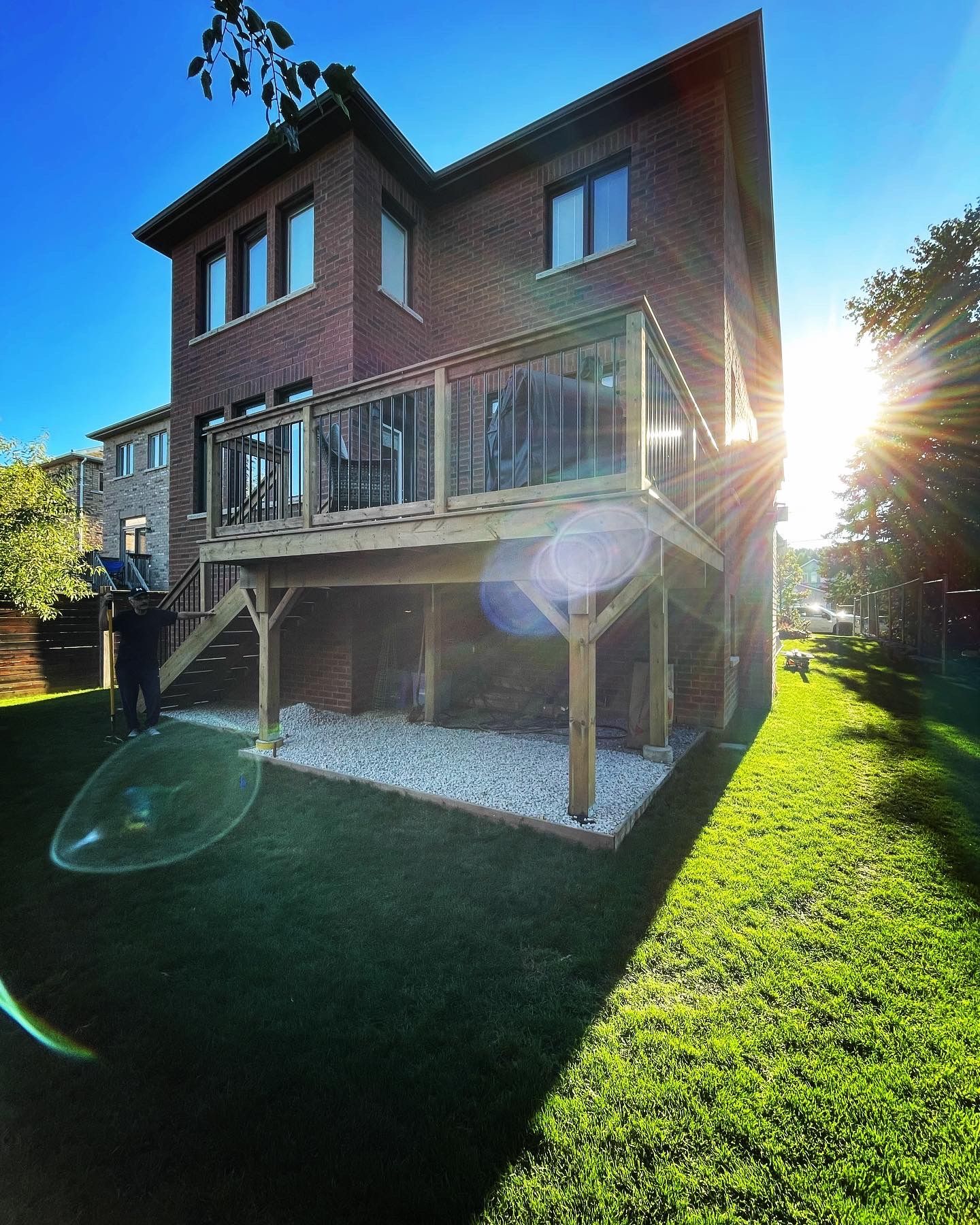 Two-story brick house with a wooden deck and glass railings, built over a gravel area. Bright sun.
