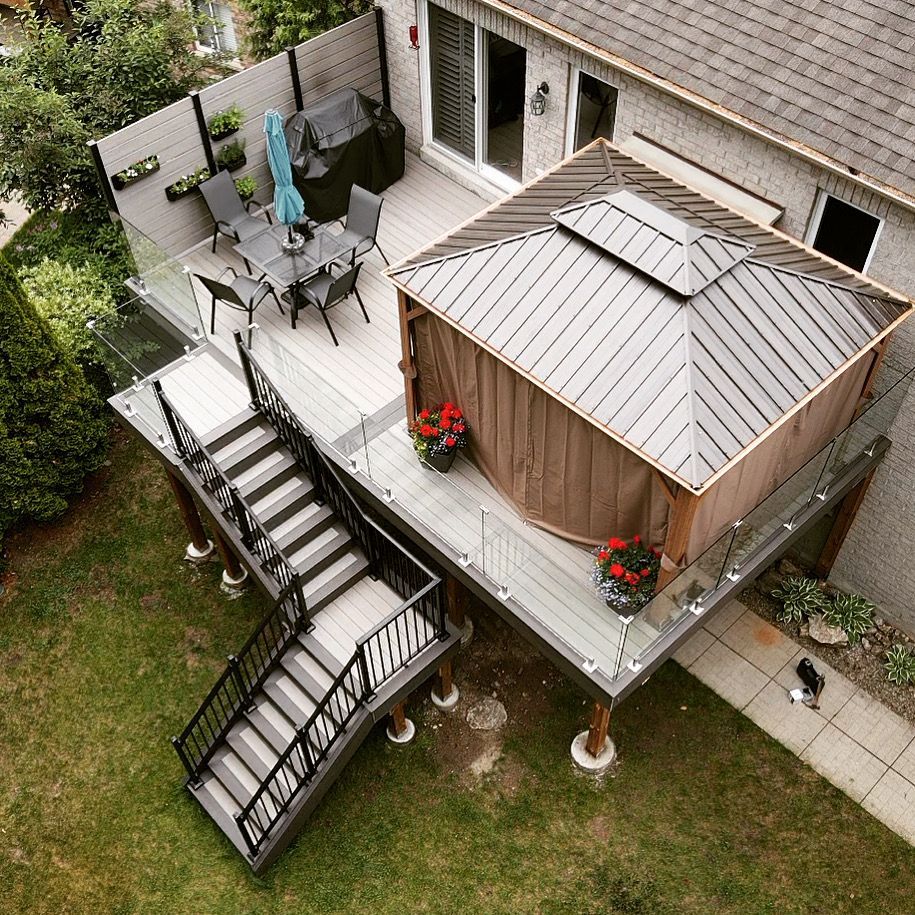 Grey second storey composite deck with a gazebo and dining area. Frameless glass railings, a large staircase with black aluminum railings