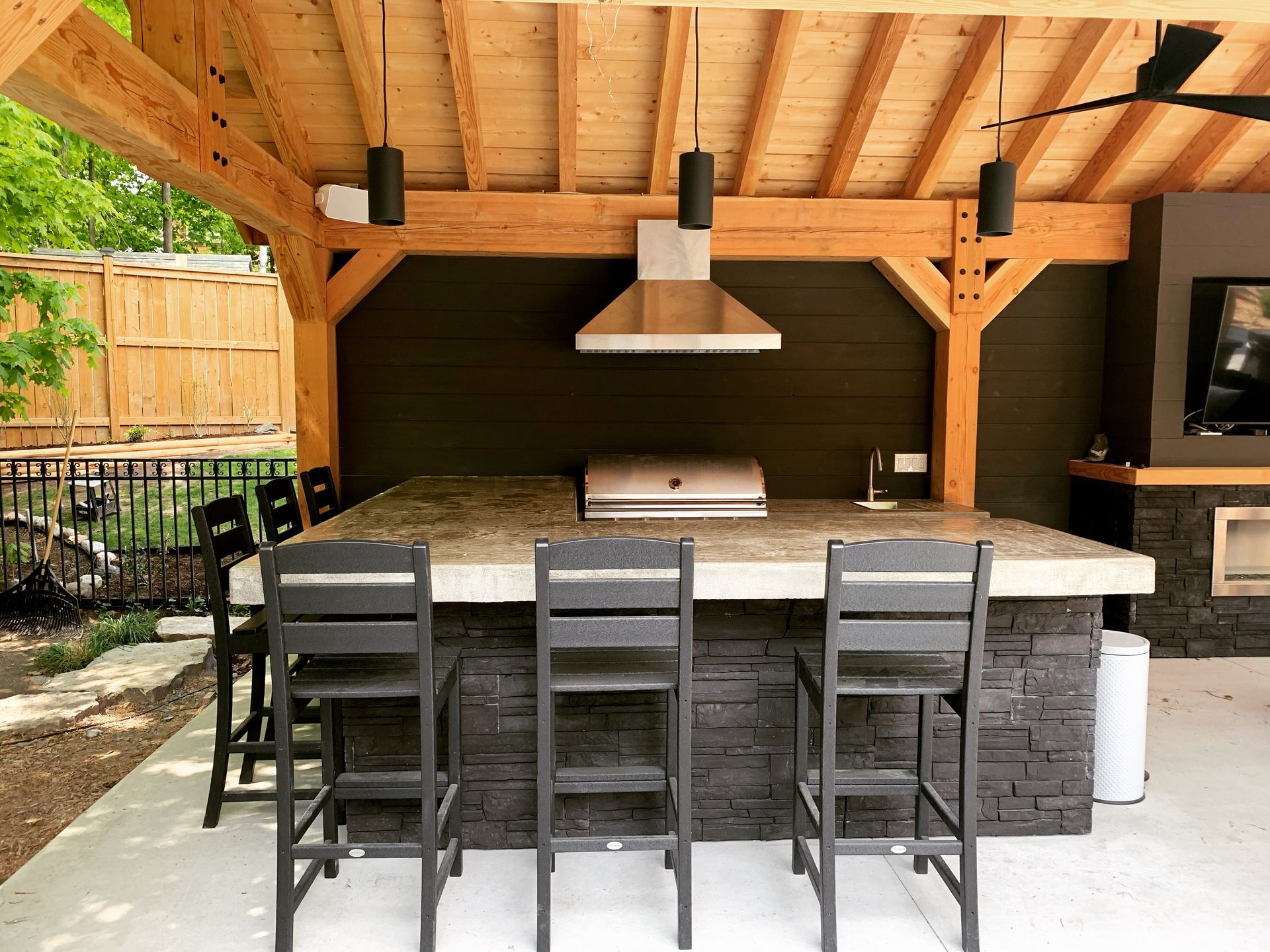 Outdoor kitchen with stone island, granite countertop, grill, and wood-framed roof. Black bar stools.