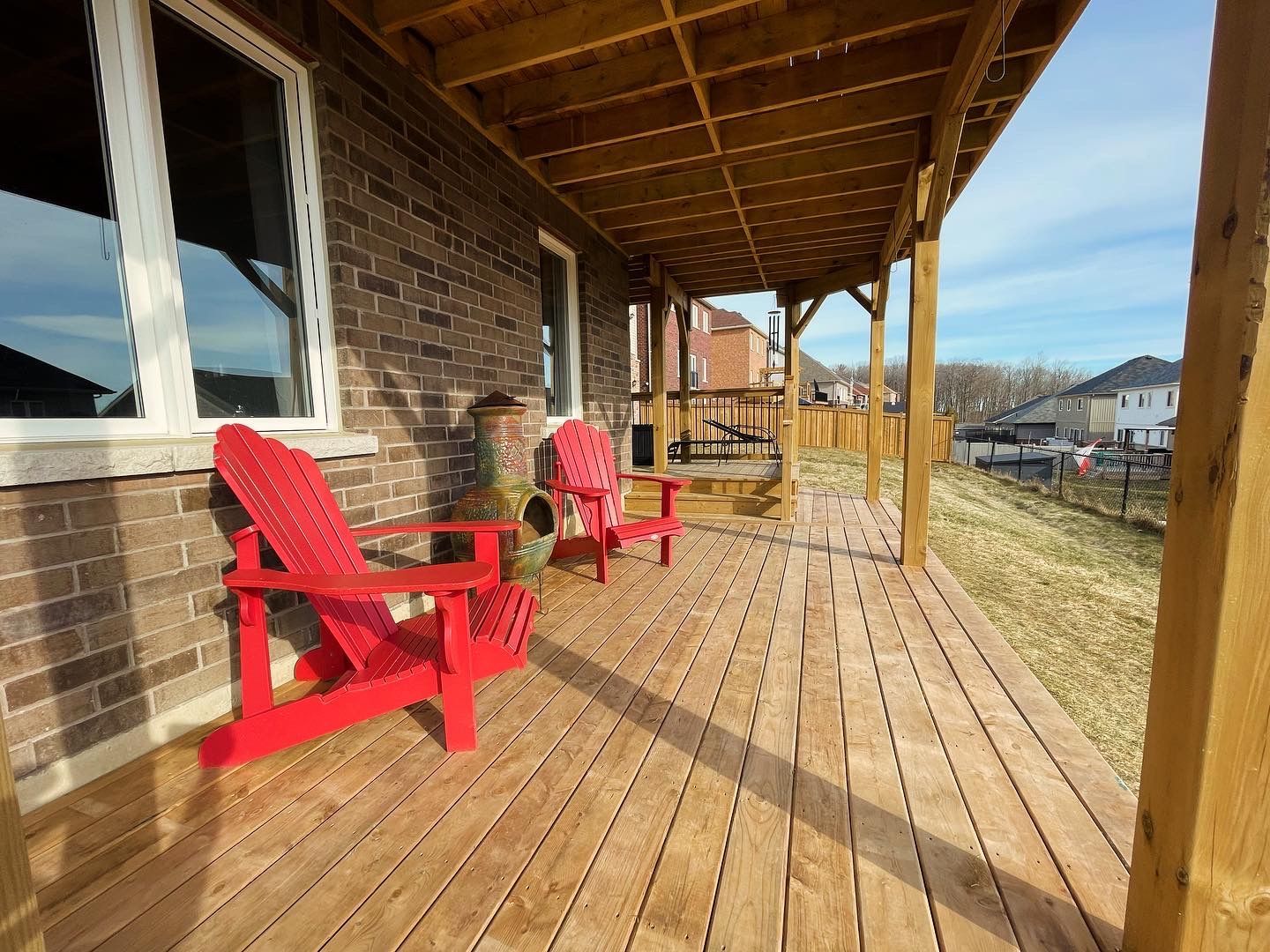 Wooden deck with two red Adirondack chairs under a covered porch; brick wall and blue sky.