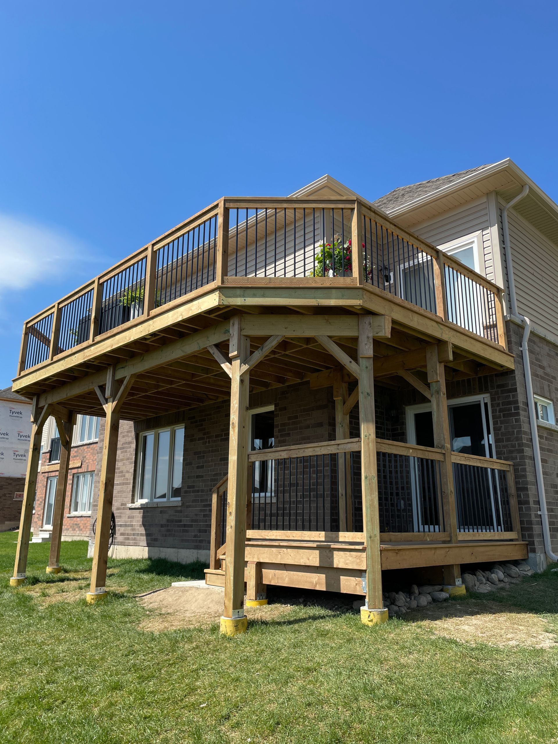 Two-story wooden deck attached to a brick house, black railings, clear sky, green grass.