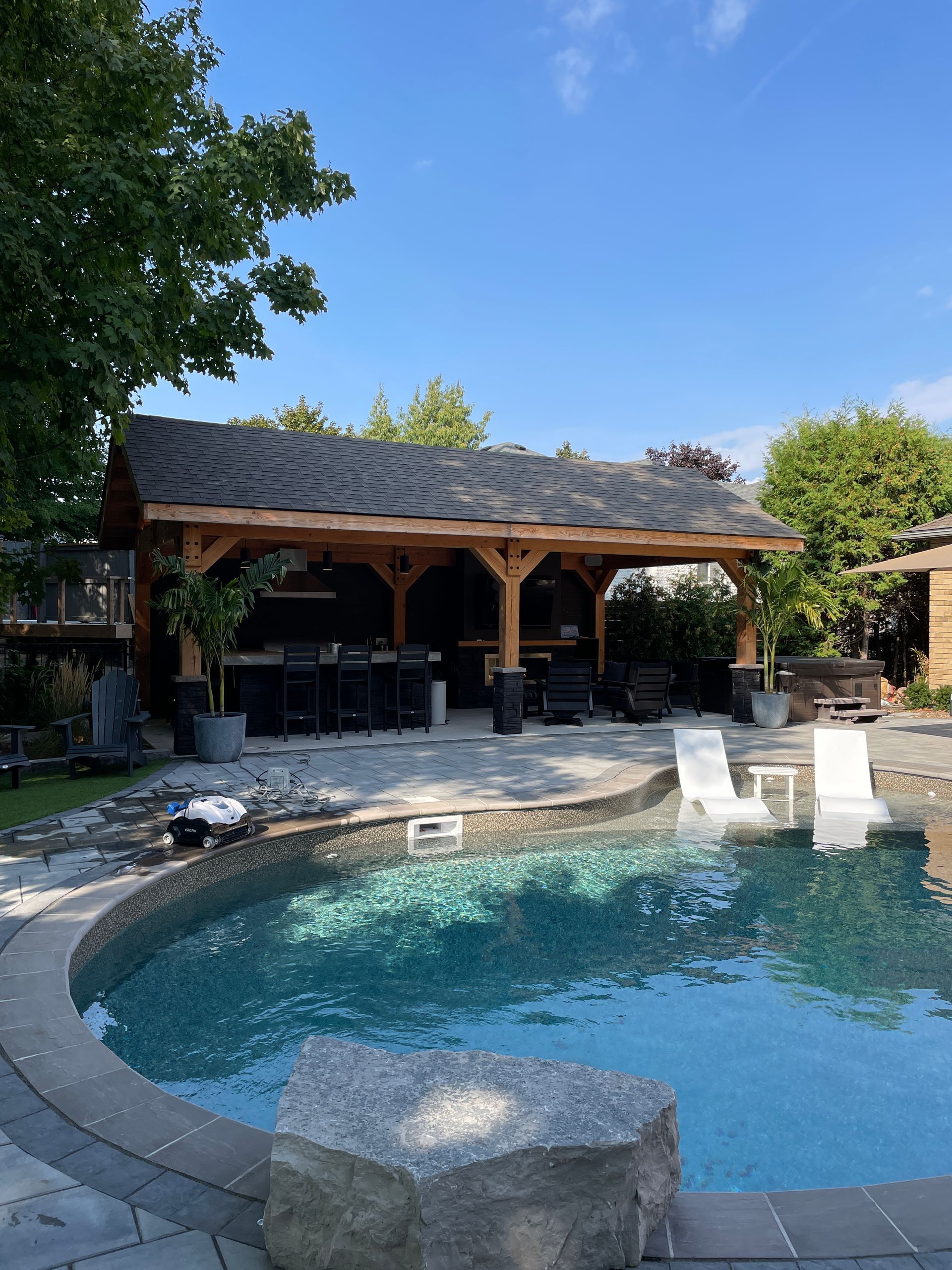 Poolside wooden pavilion with seating, next to a pool under a blue sky.