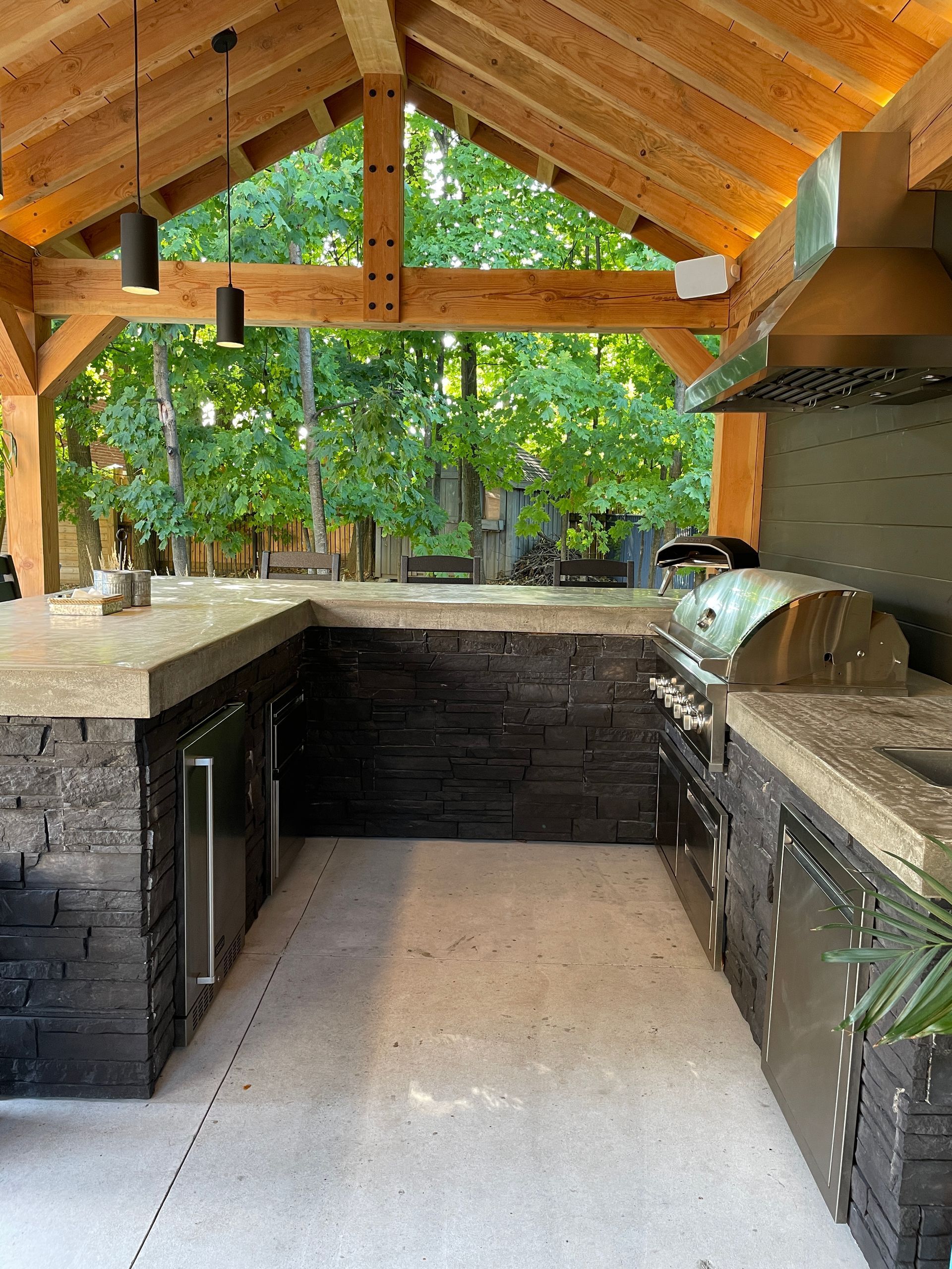 Outdoor kitchen with stone facade, under a wooden roof. Includes grill, countertop, and appliances.