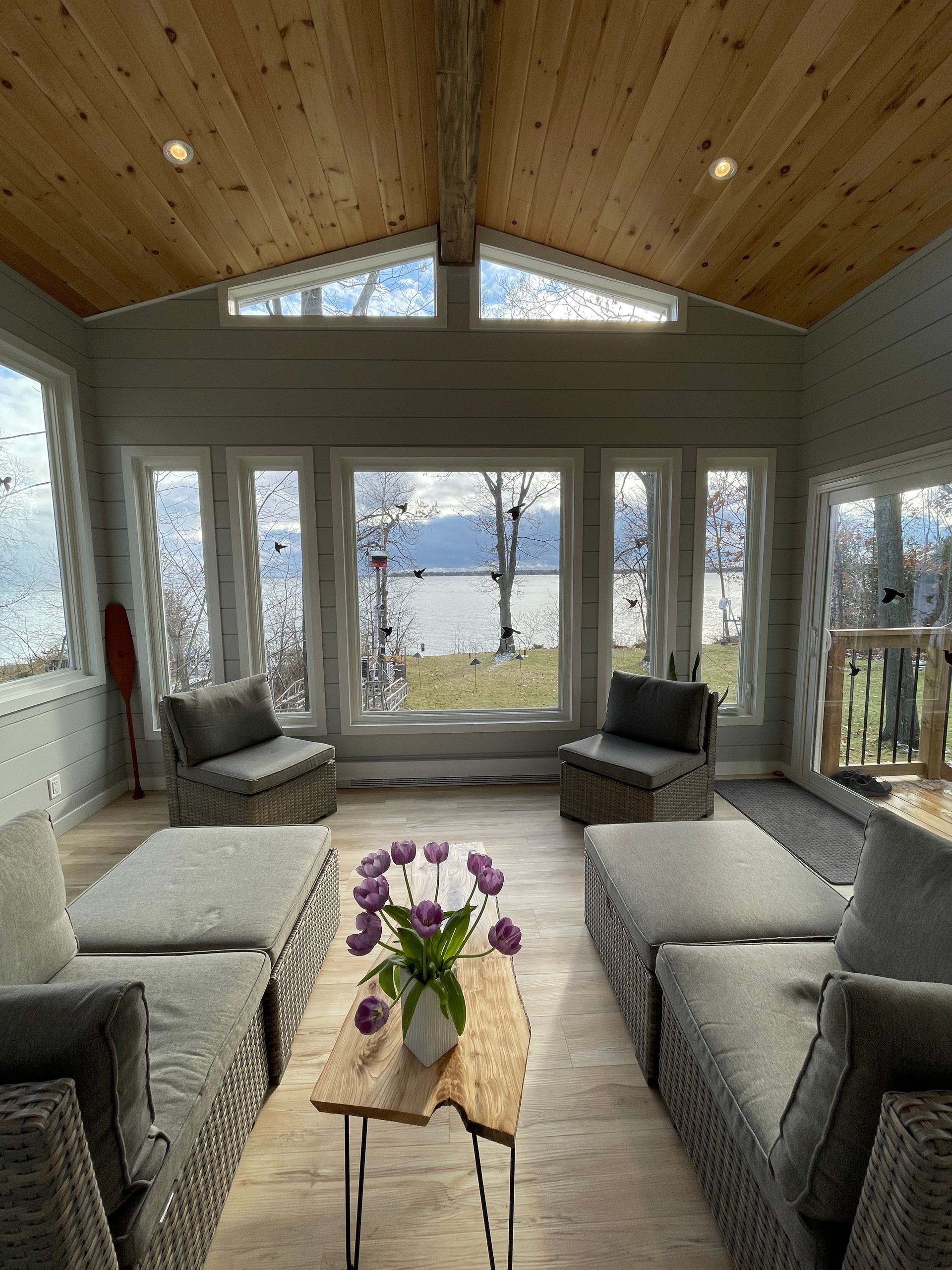 Sunroom with grey seating, wood ceiling, and large windows overlooking water and trees.