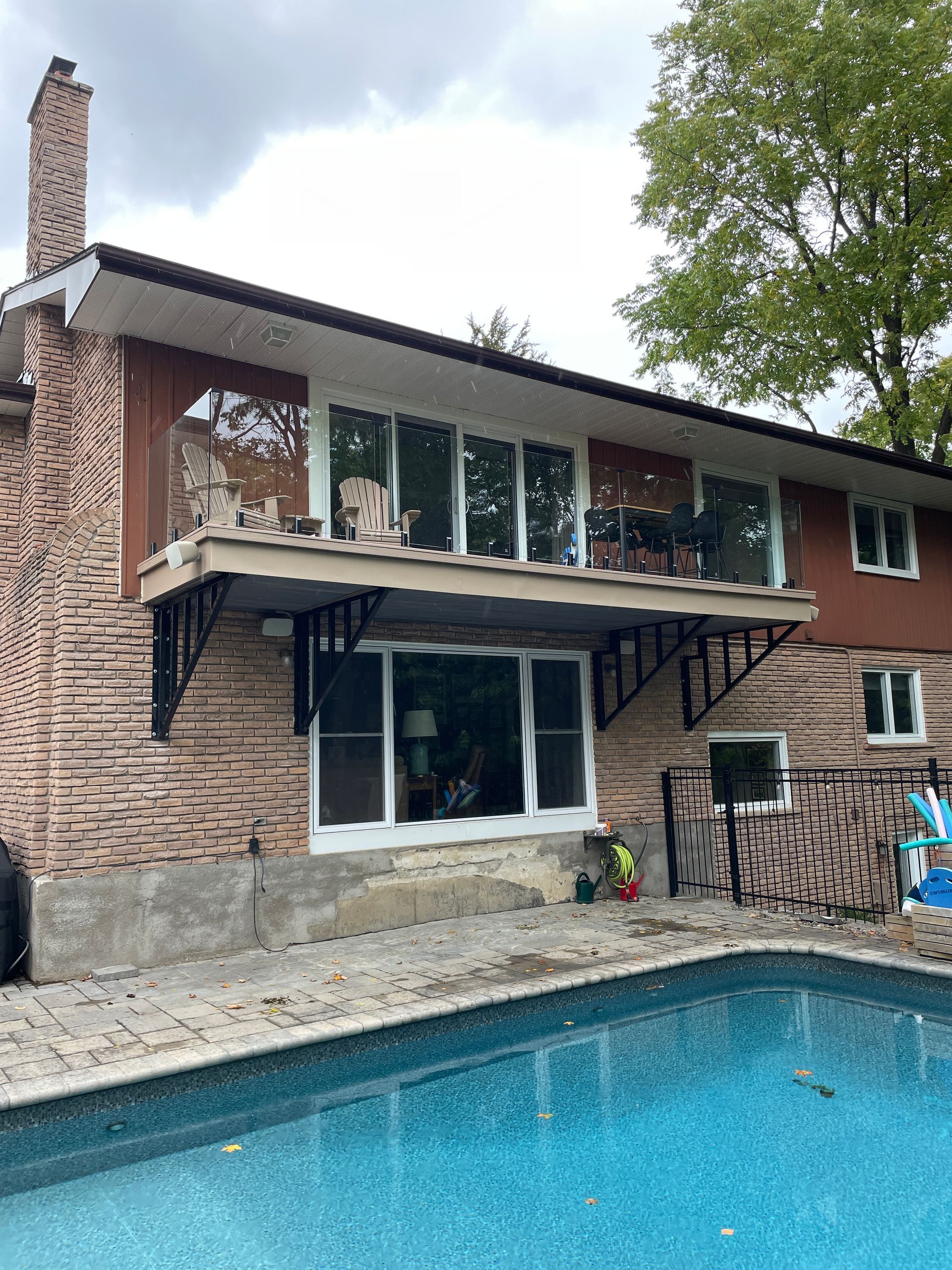 Back of a house with a pool. Balcony with black brackets, brick exterior, and large windows. Blue pool in the foreground.