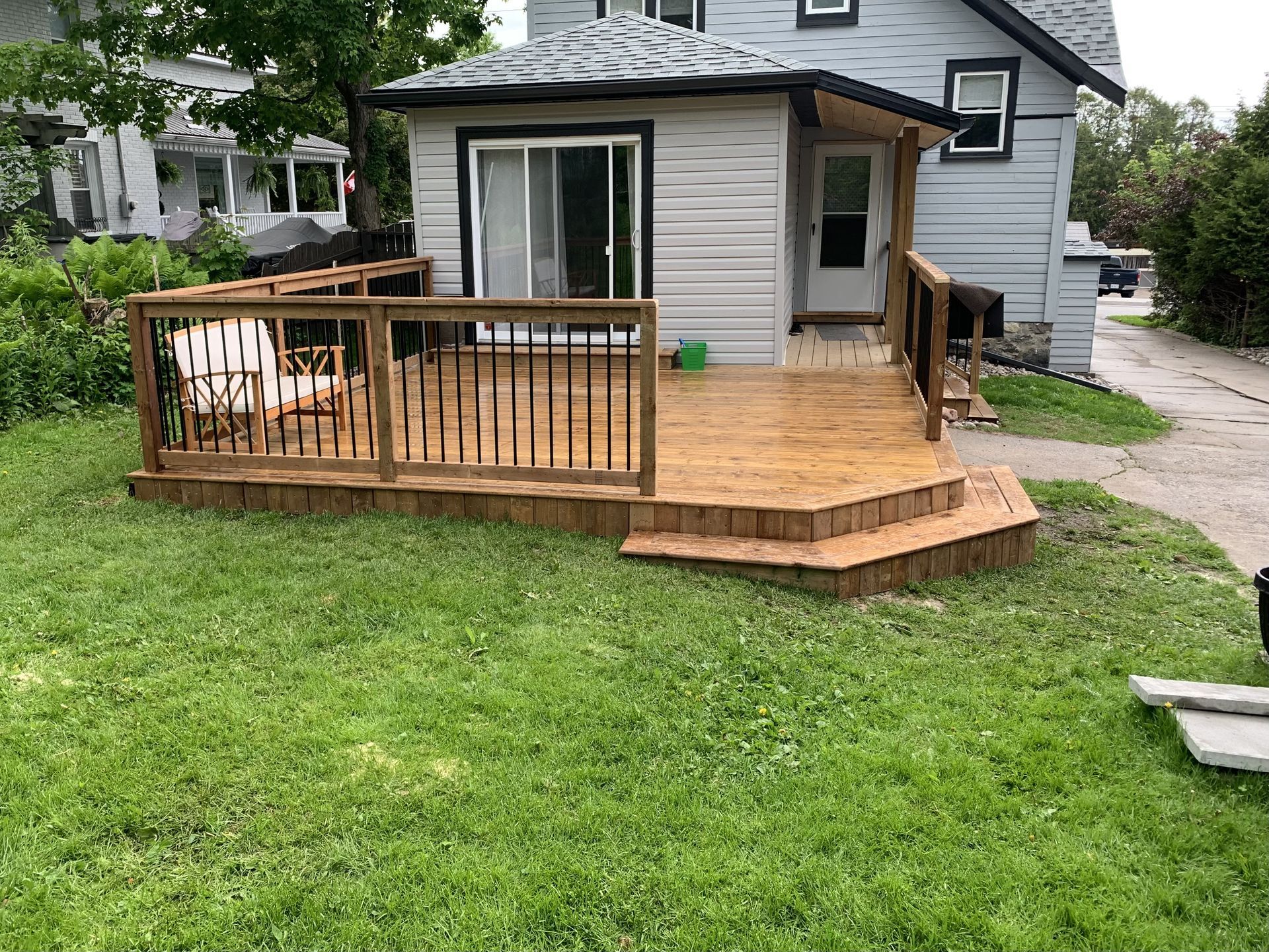 Wooden deck with railings and steps in a yard, connected to a light gray house with a sliding door and a white door.