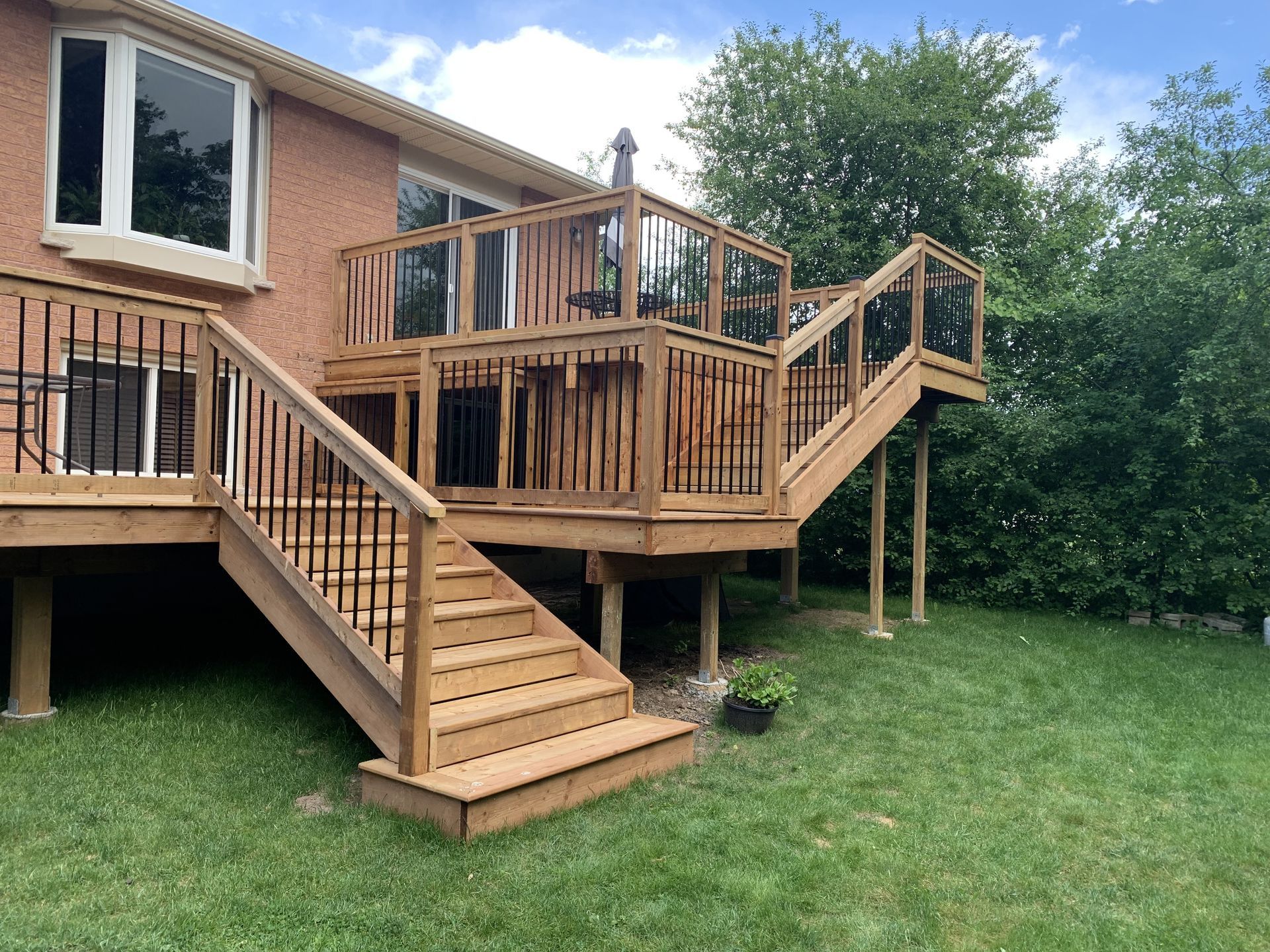 Wooden deck with stairs, glass and black metal railing, against a brick house, on a grassy lawn.