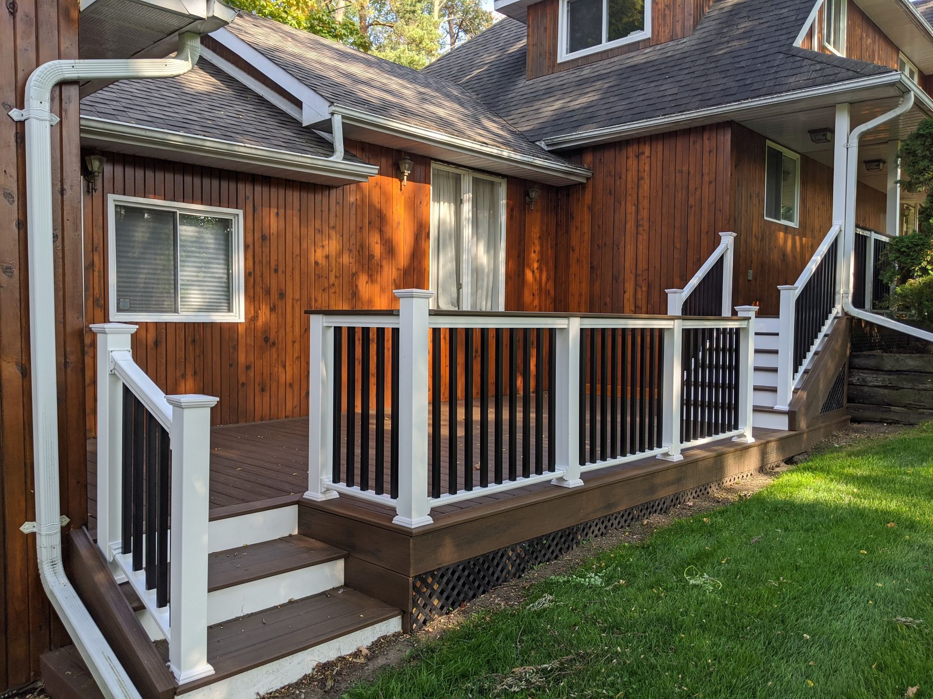 Wooden deck with white and black railings, brown siding, and stairs leading to a house. Green grass surrounds it.