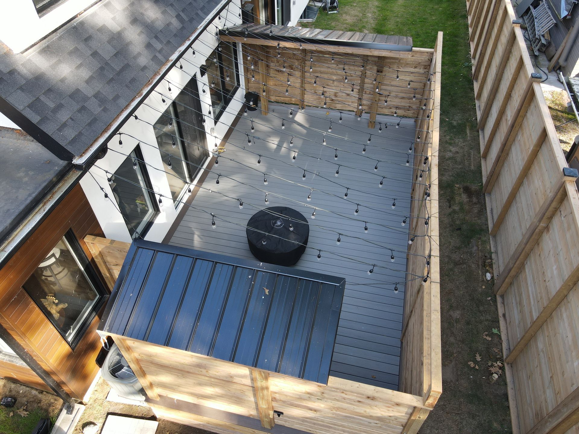 Aerial view of a backyard deck with a fire pit, wooden fencing, and string lights.