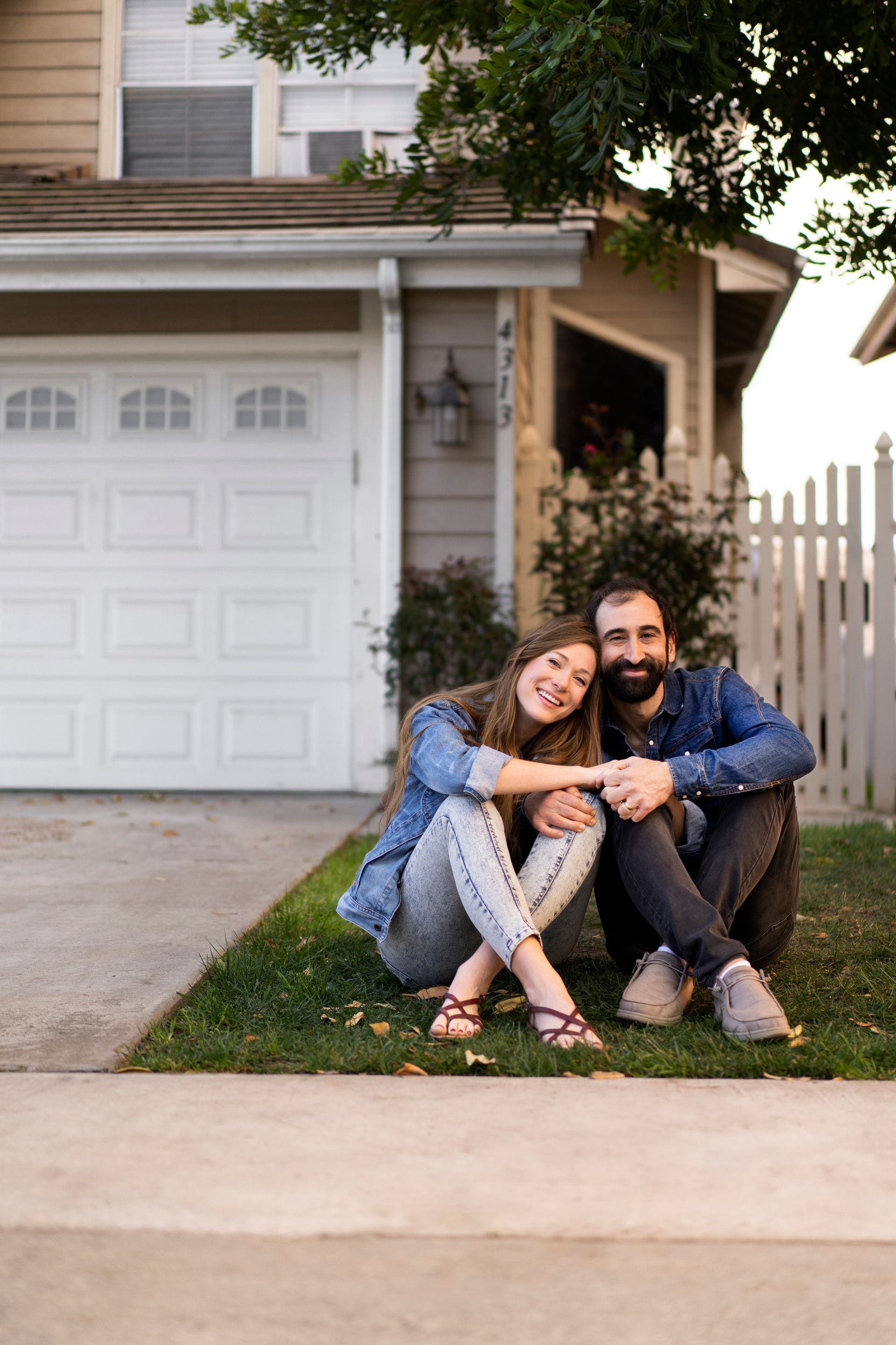 A smiling couple sits on the grass in front of their house. The woman leans on the man's shoulder, arms crossed, and they are both wearing denim jackets.