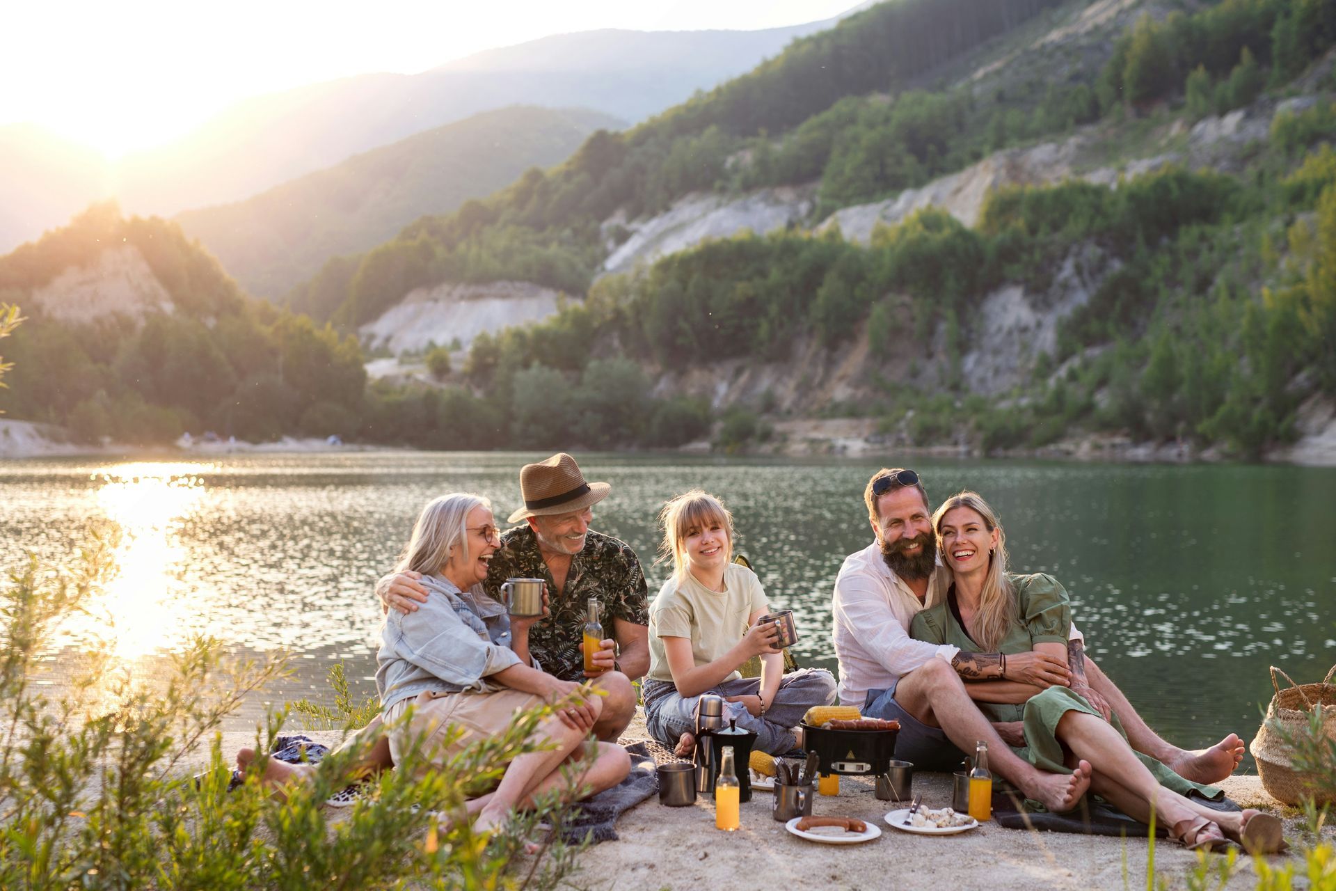 Family picnic by a lake. People of different ages seated together, enjoying food and drinks, with a scenic mountain backdrop.