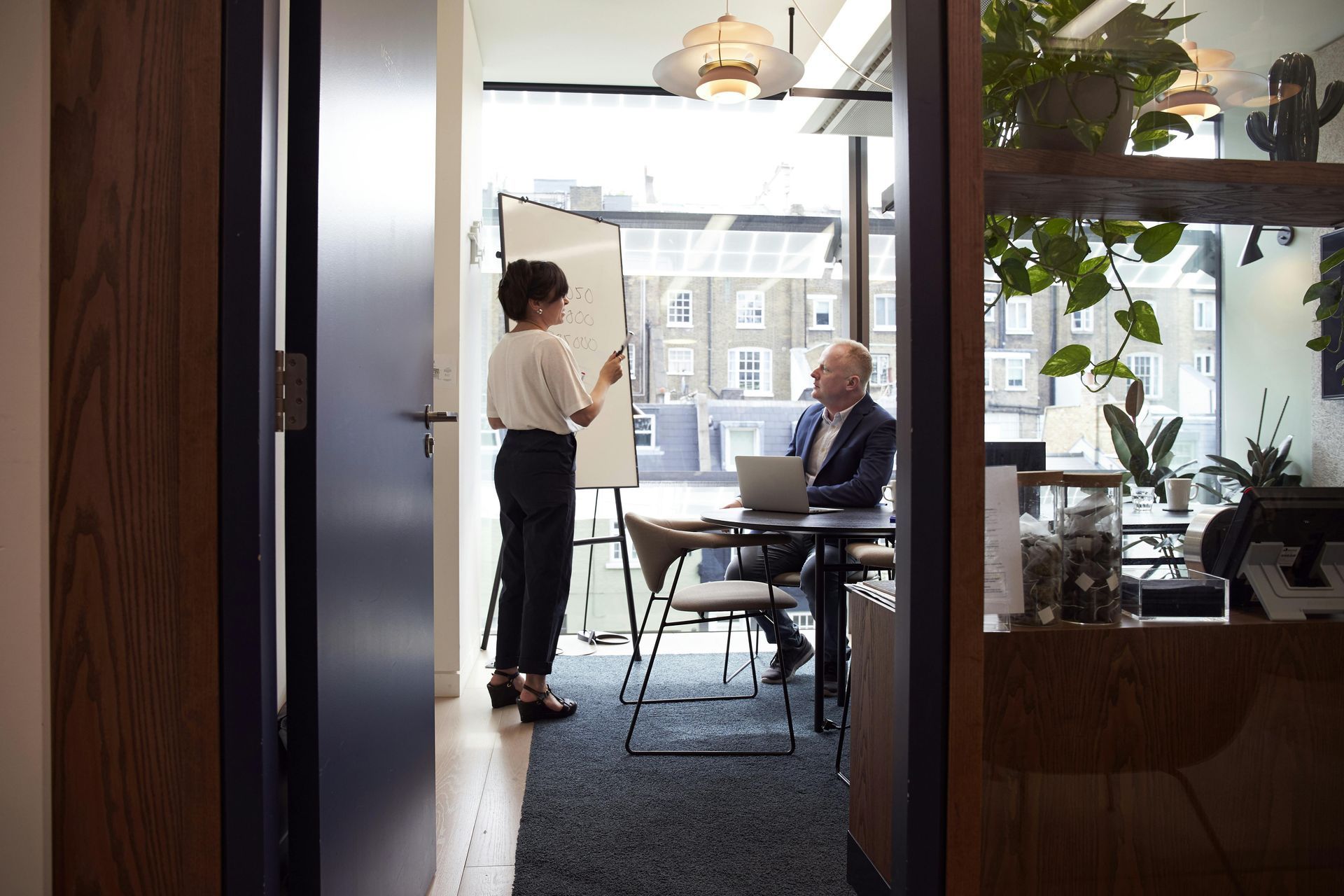 Woman presenting to a man in an office. The woman stands at a whiteboard, pointing. The man sits at a desk, looking at a laptop.