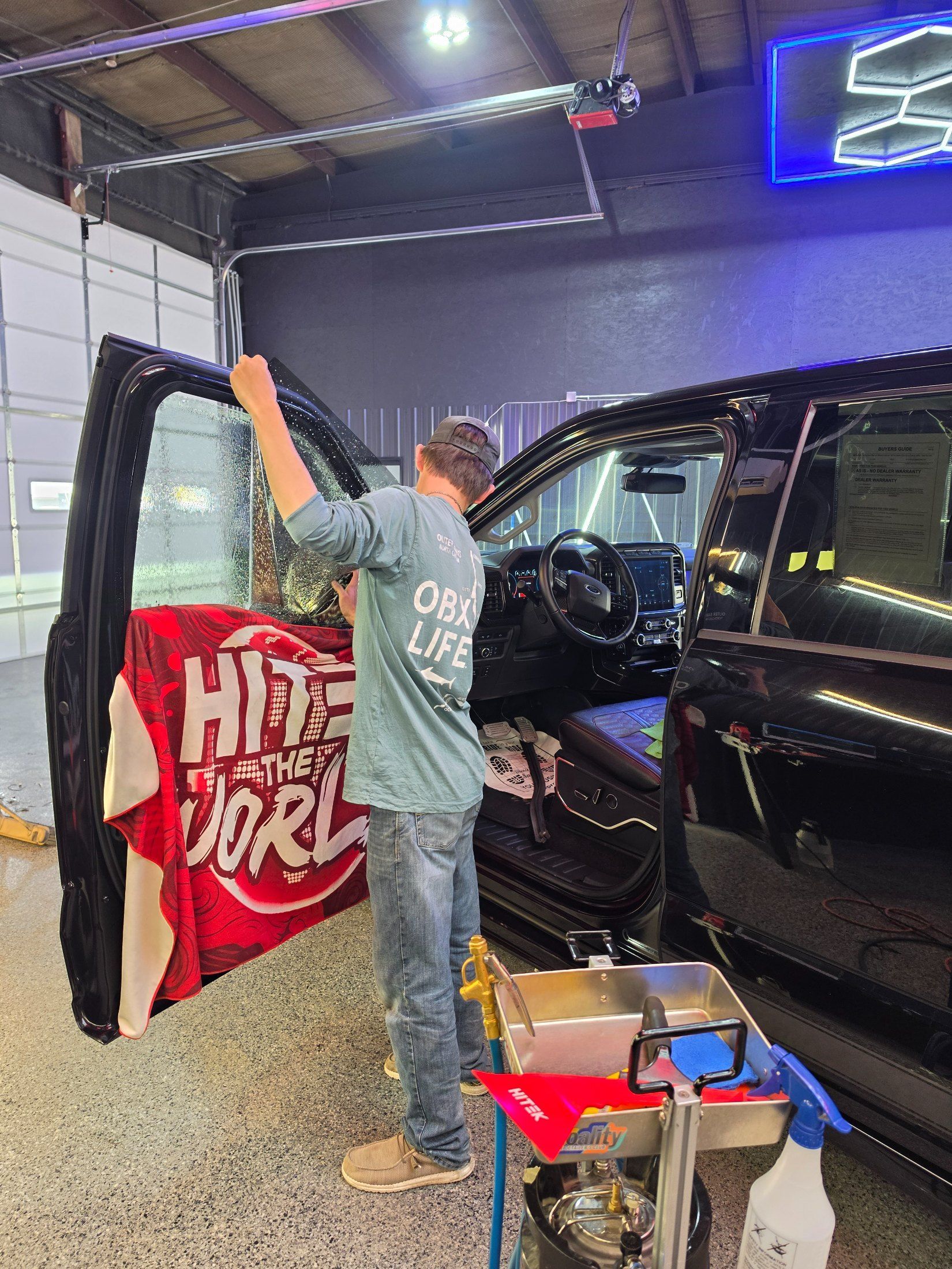 Person in a workshop raises arms beside a black vehicle and red sign; tools and boxes nearby.