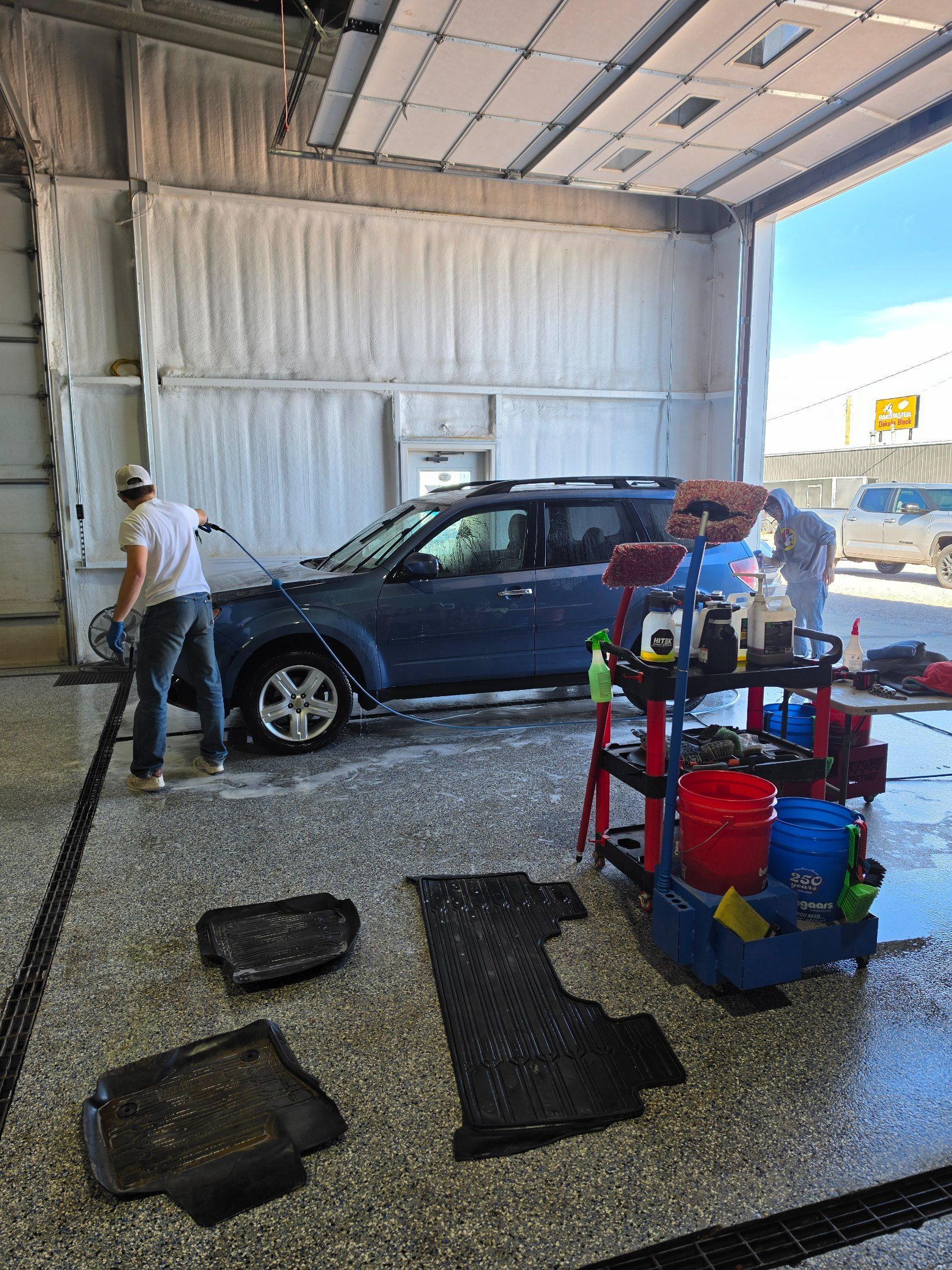 Blue sedan in a garage bay with two workers and a cleaning cart on a wet floor.