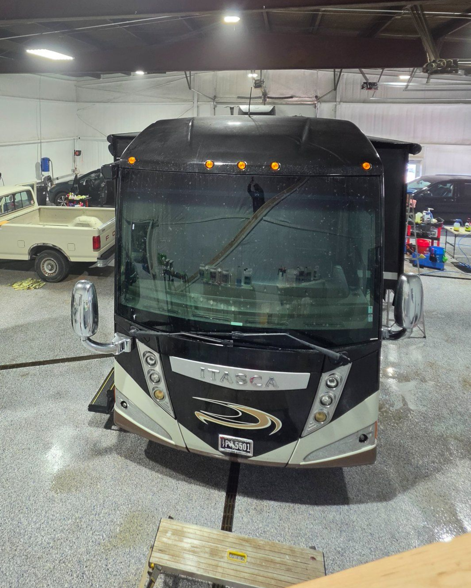 Front view of a black and silver tour bus inside a garage, with lights on and wet floor.