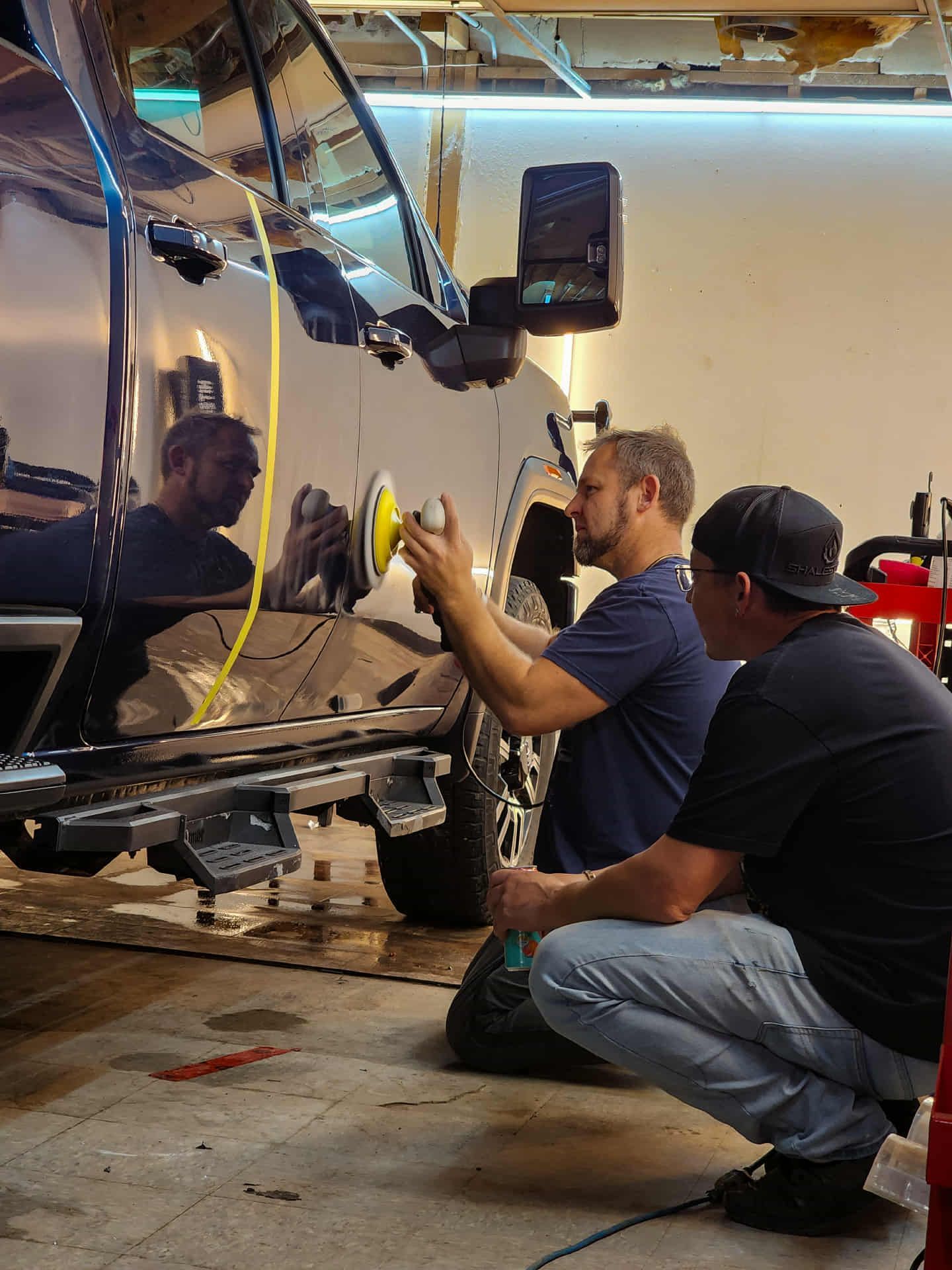 Two men polishing a black vehicle. One uses a buffer, the other watches. Inside a garage.