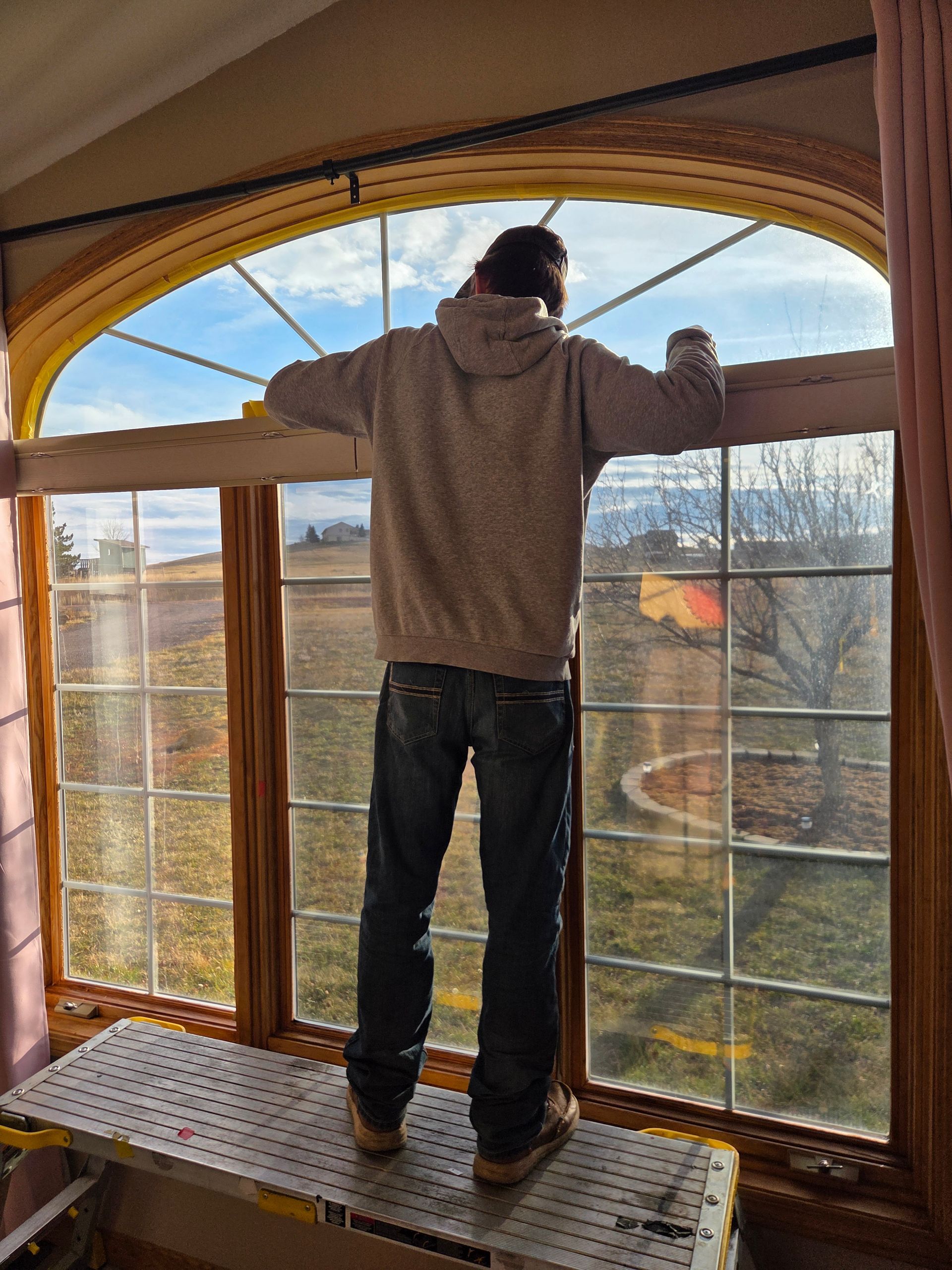 Person on a platform cleaning a large arched window with a view of a cloudy sky and field.