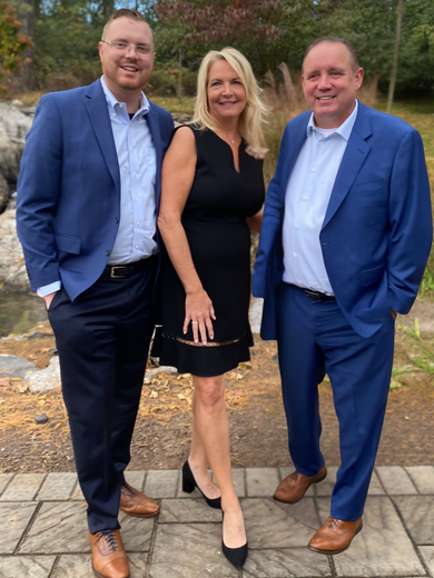 Three people posing outdoors. Woman in black dress between two men in blue suits.
