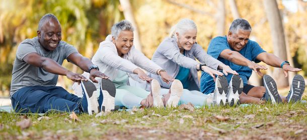 Four older adults stretching their legs while sitting on grass in a park, smiling.