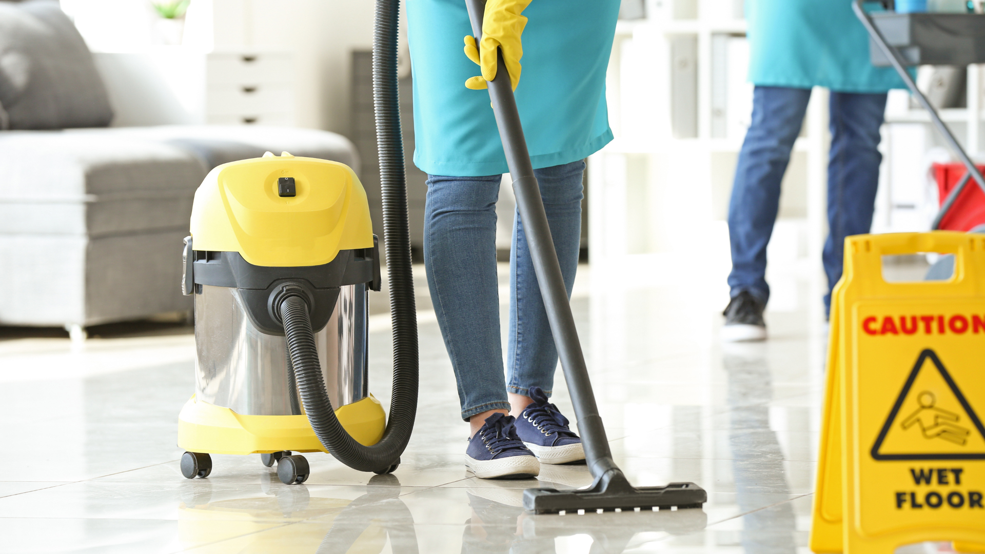 Person vacuuming shiny floor near caution sign and vacuum cleaner.