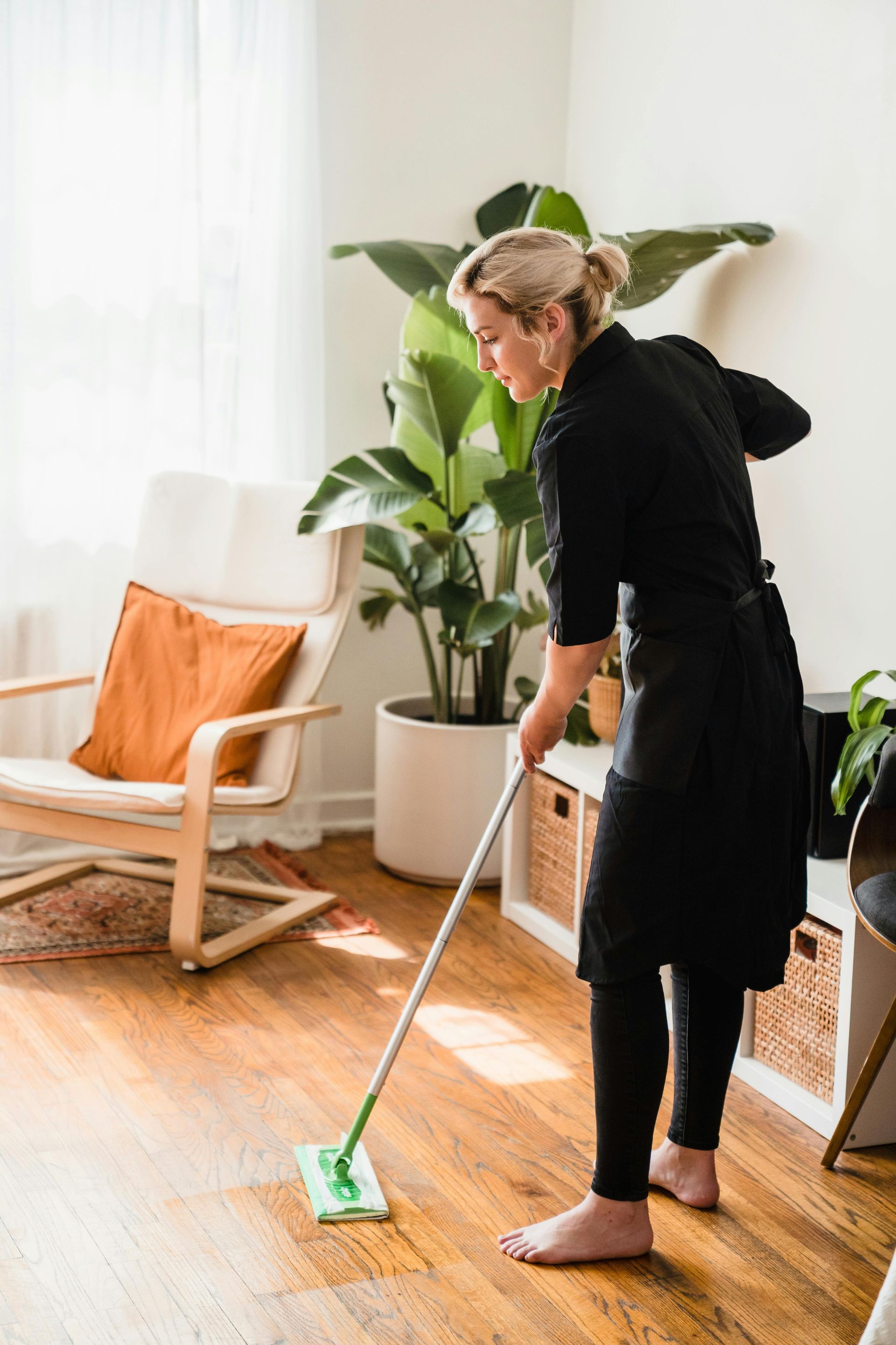 Woman mopping wooden floor in a brightly lit room with a potted plant and a chair.