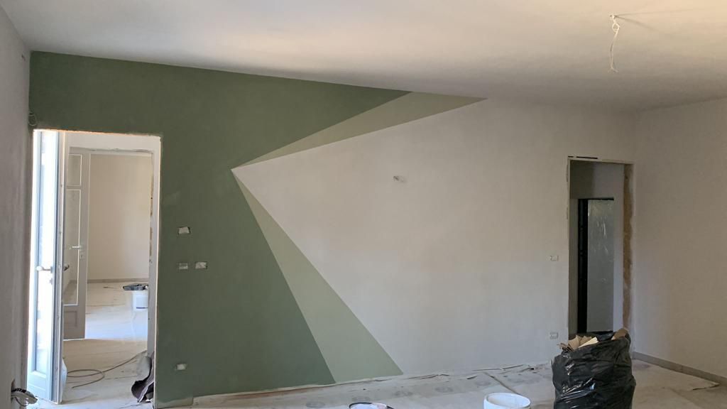 Interno di una stanza con parete decorativa dipinta con motivi geometrici: triangoli verdi e bianchi. Porte aperte, non rifinite.