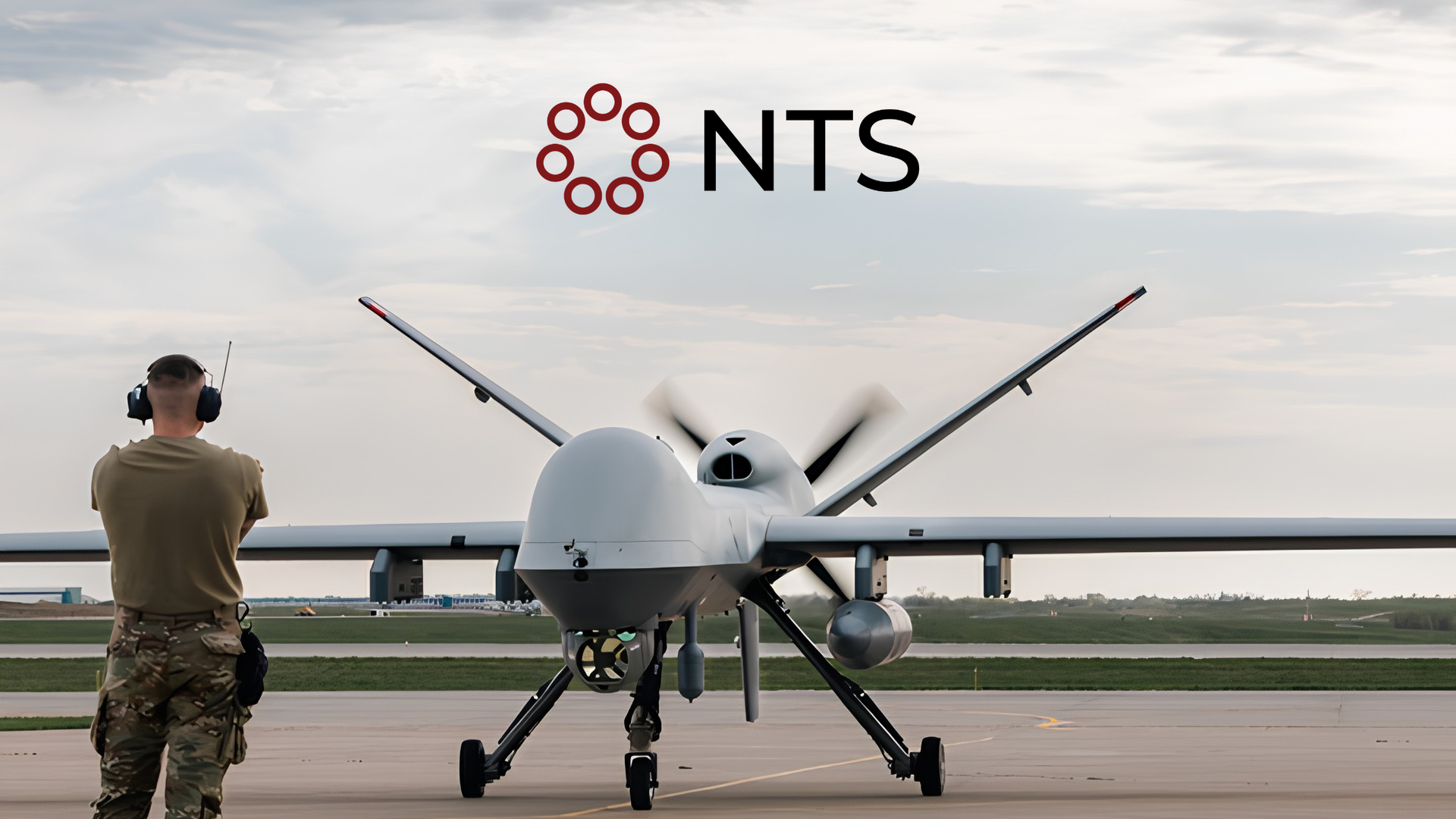 Military drone on runway with a person standing beside it under an NTS logo