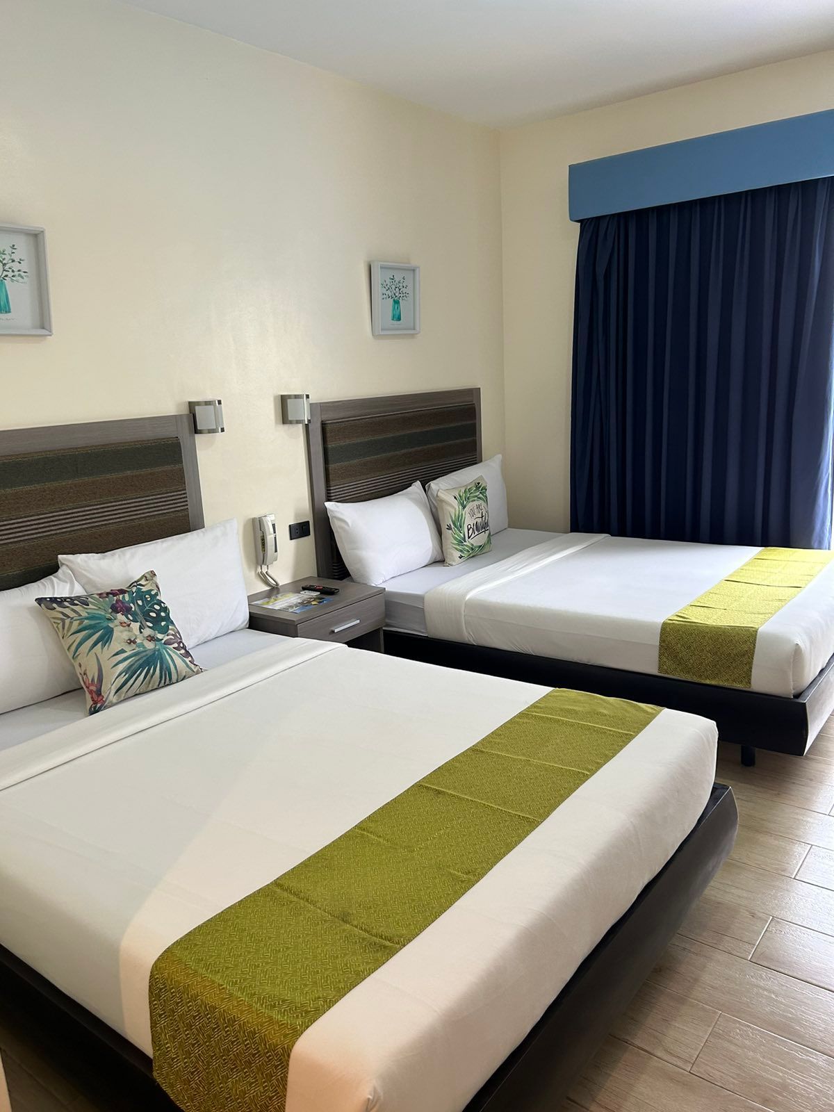 Two queen beds in a hotel room with green bed runners, white linens, and a blue curtain.