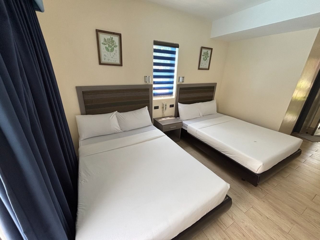 Two beds in a hotel room with blue curtains, two framed pictures on the wall, and a window with a blind.