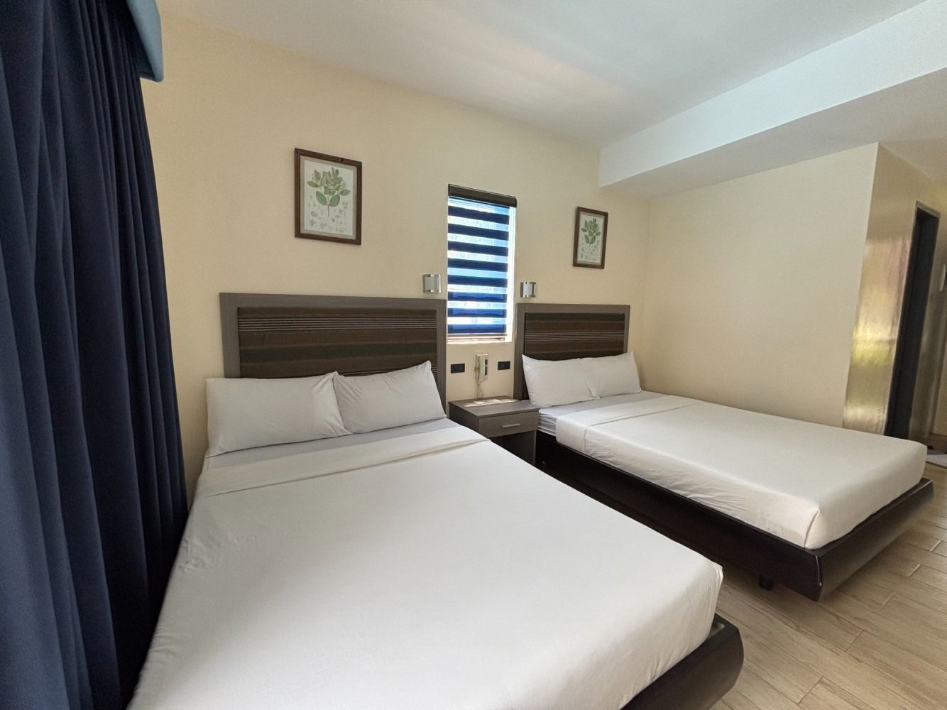 A hotel room with two beds, light walls, blue curtains, and framed botanical prints.