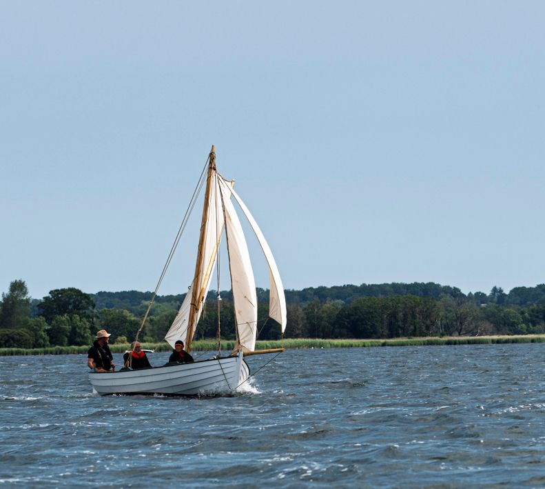 Sejlbåd med tre personer, der sejler på havet, hvide sejl, solskinsdag.