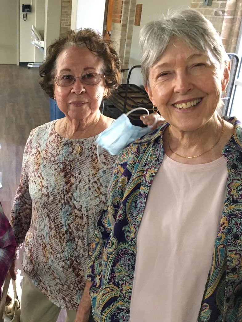 Two smiling people stand indoors, one wearing a patterned top and the other in a light shirt with an open print jacket.