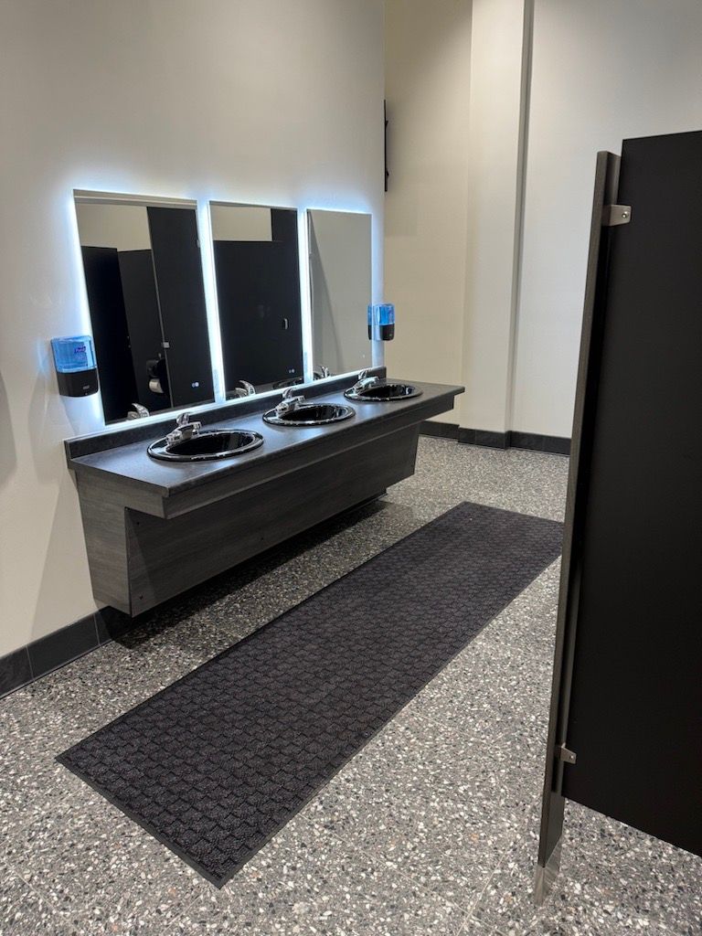 Public restroom with three sinks, mirrors, soap dispensers, and a black stall door on tiled floor.