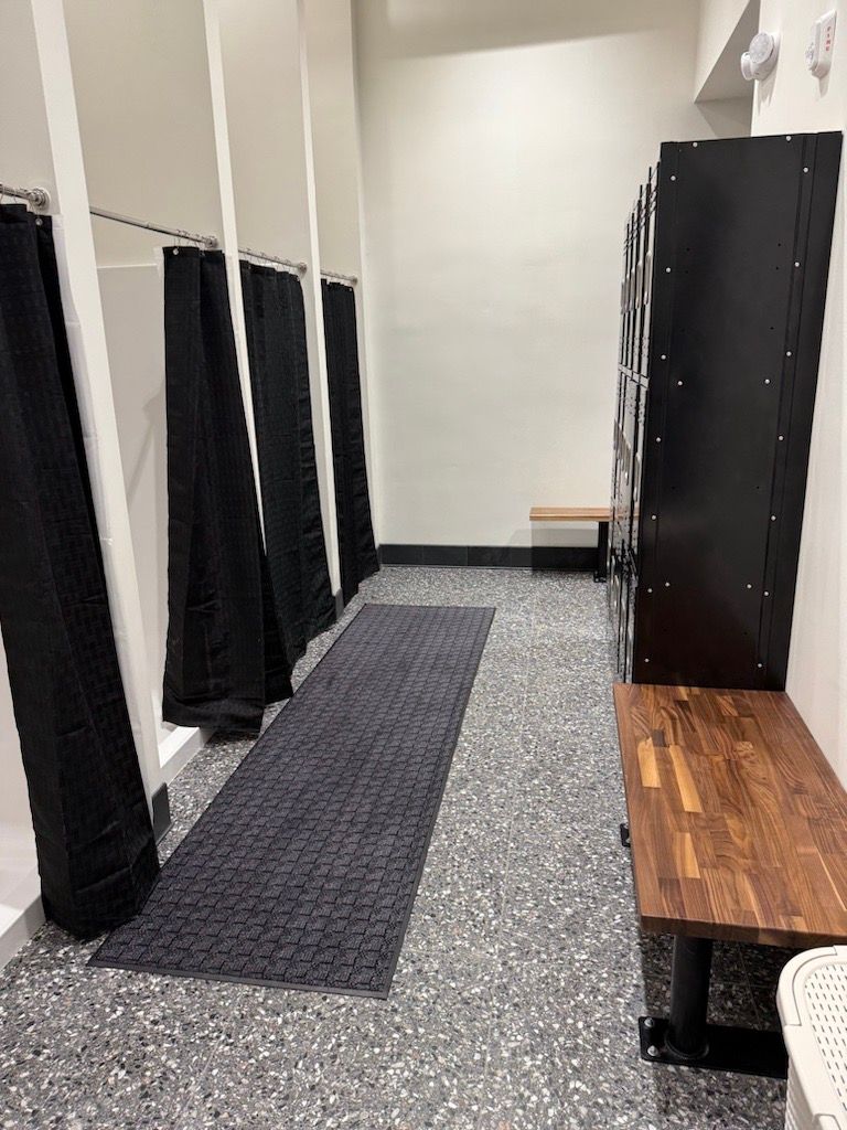 Locker room with black stalls, gray floor mats, wooden bench, and a white wall.